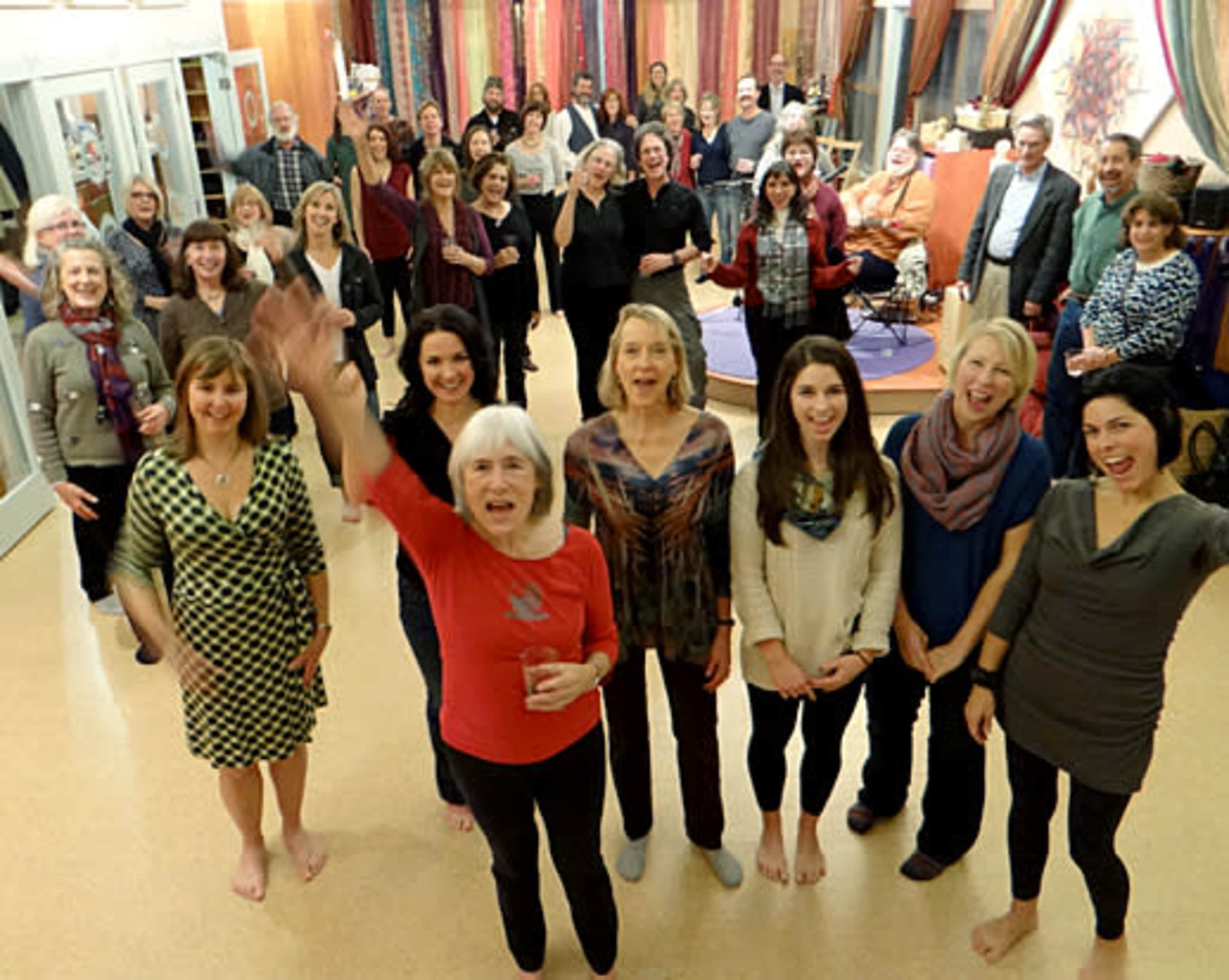 A diverse group of people stands together in a brightly lit room, some waving and smiling at the camera.