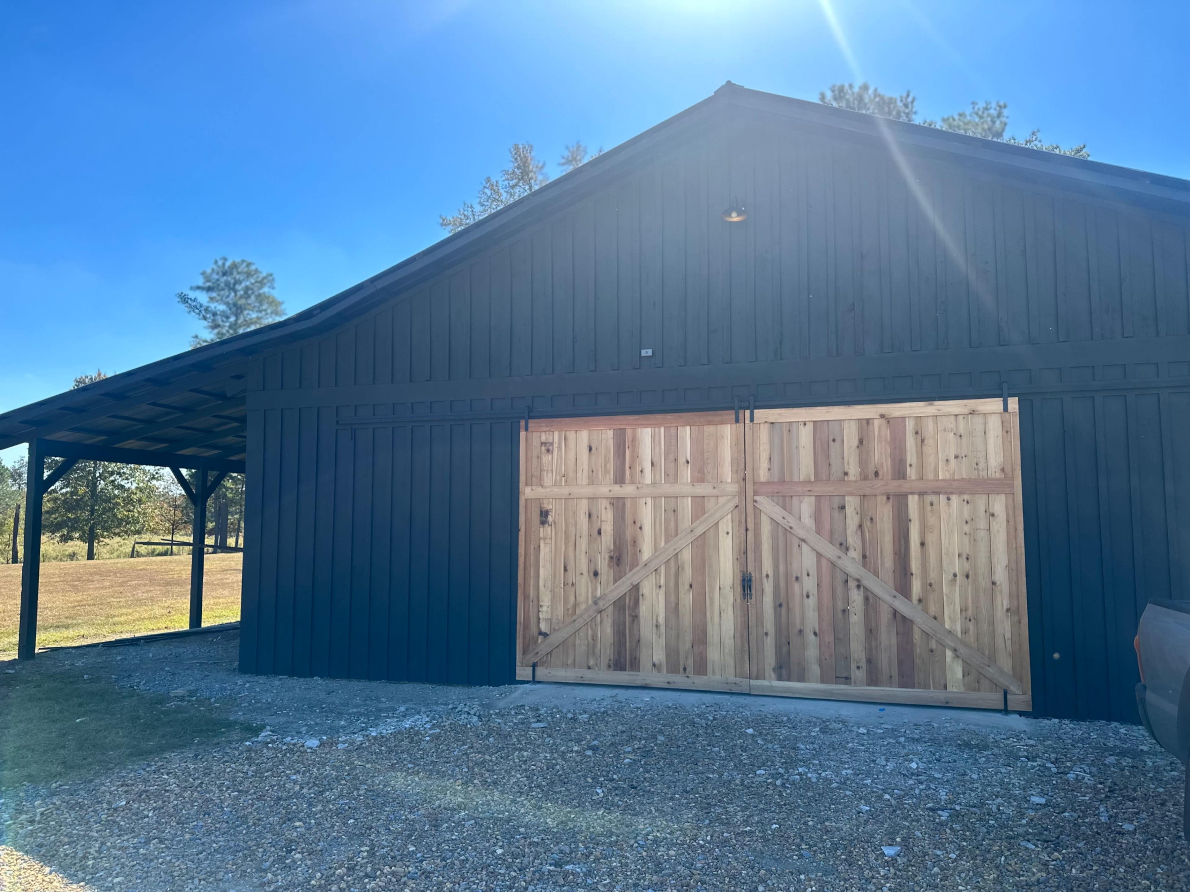 The image shows a dark wooden barn with a large wooden double door and a gravel pathway in front of it.