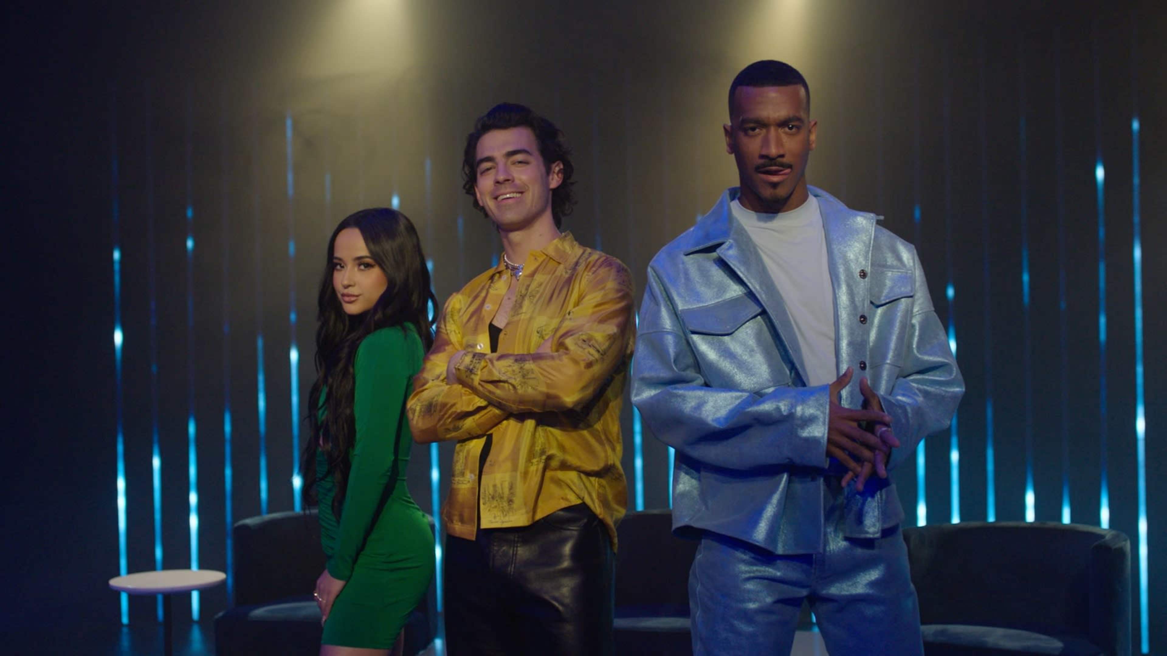 Three individuals pose confidently in front of a backdrop of vertical blue lights, with one wearing a green dress, another in a yellow patterned shirt, and the last in a shiny silver suit.