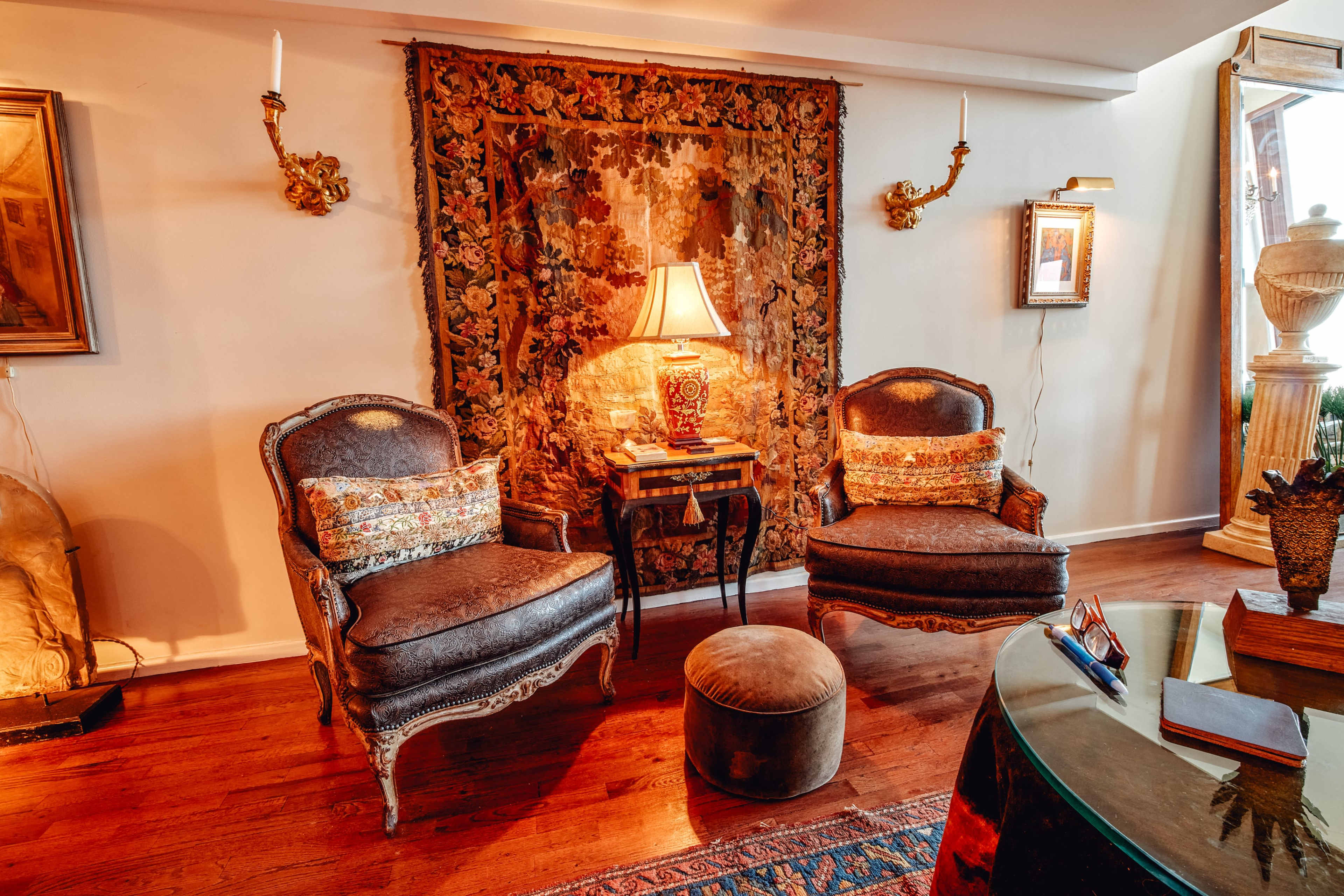 The image shows two upholstered chairs with decorative pillows positioned beside a small table, illuminated by a lamp, against a backdrop of a colorful tapestry on the wall.