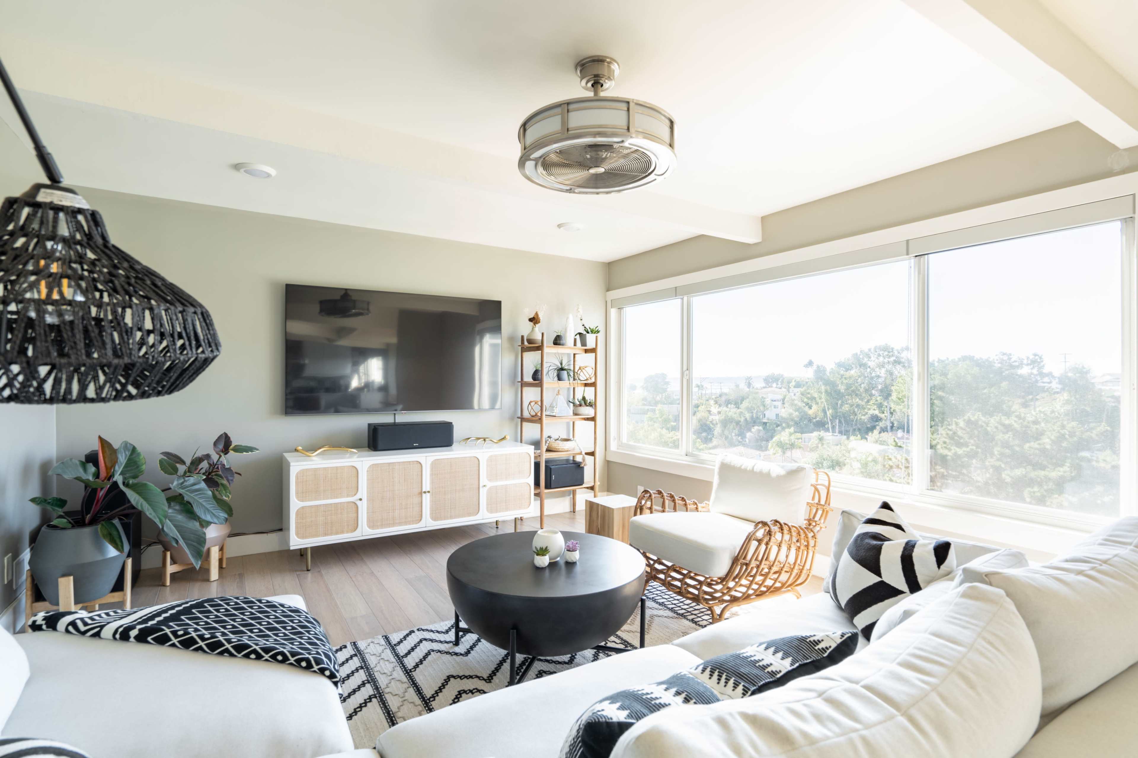 The image shows a modern living room featuring a light-colored sofa, a round black coffee table, and large windows with a view of greenery outside.