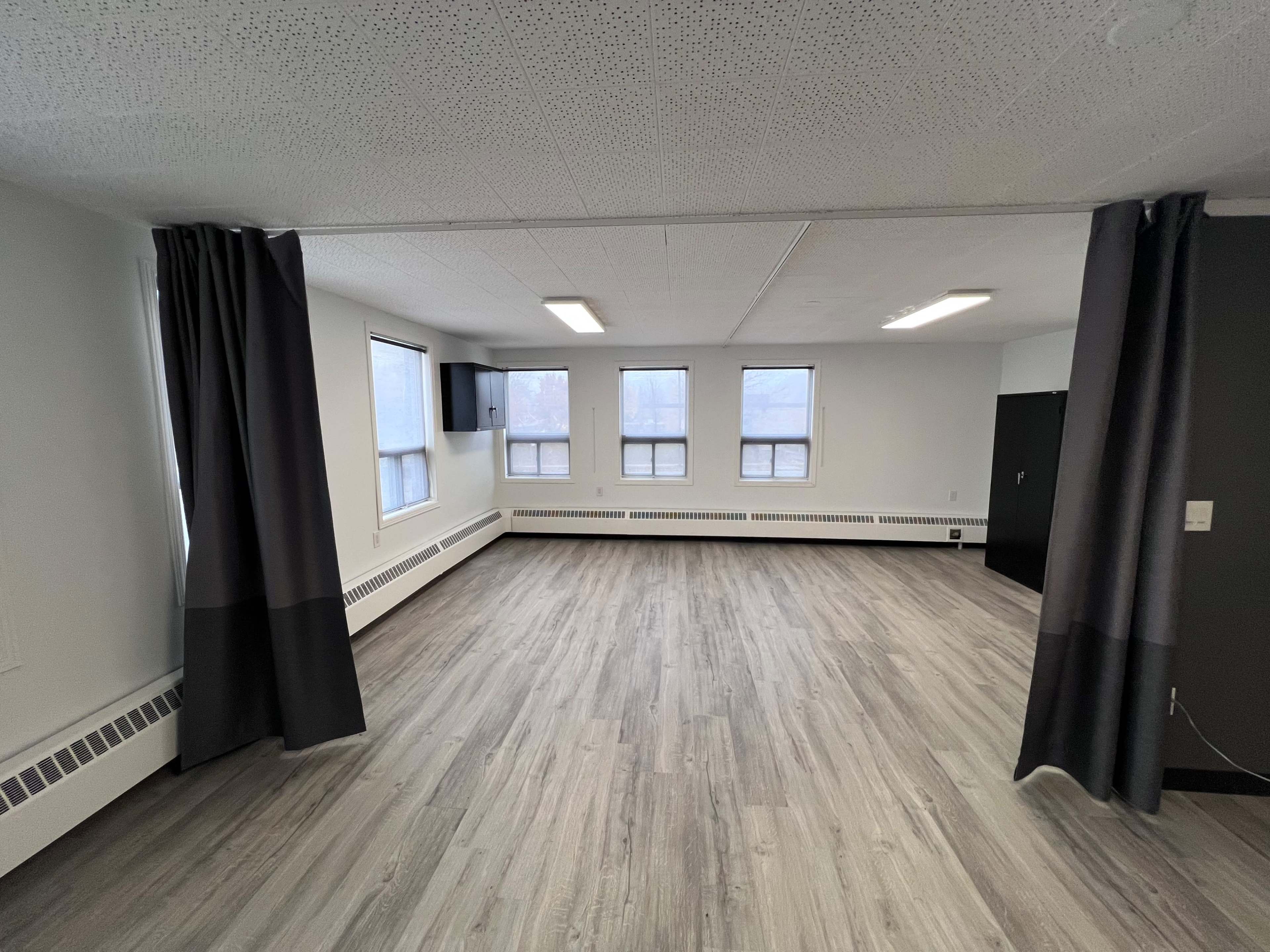 An unfurnished, open room with light wood flooring, large windows, and gray curtains on one side.