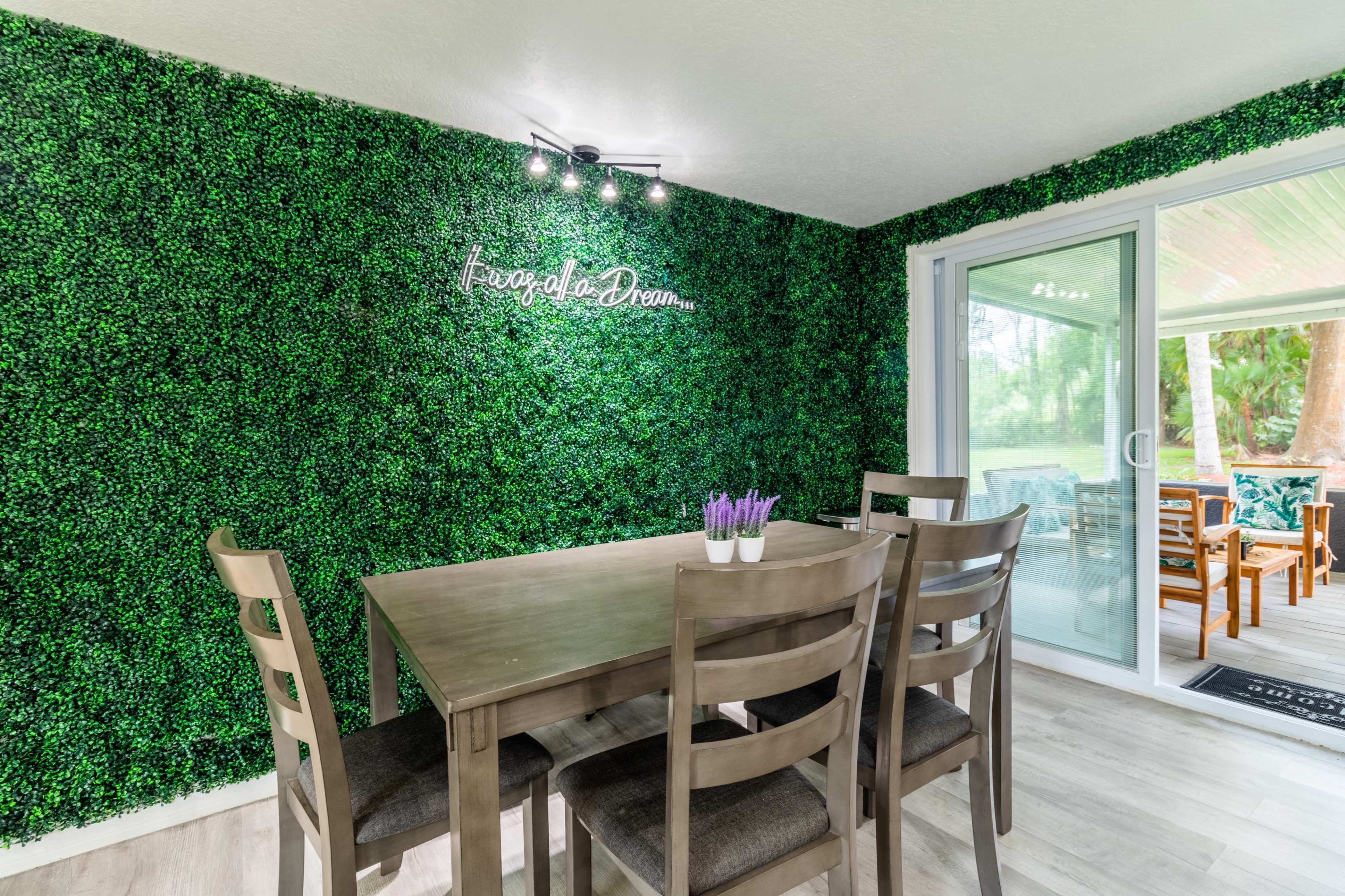 A dining area features a wooden table surrounded by chairs, set against a wall covered in artificial greenery.