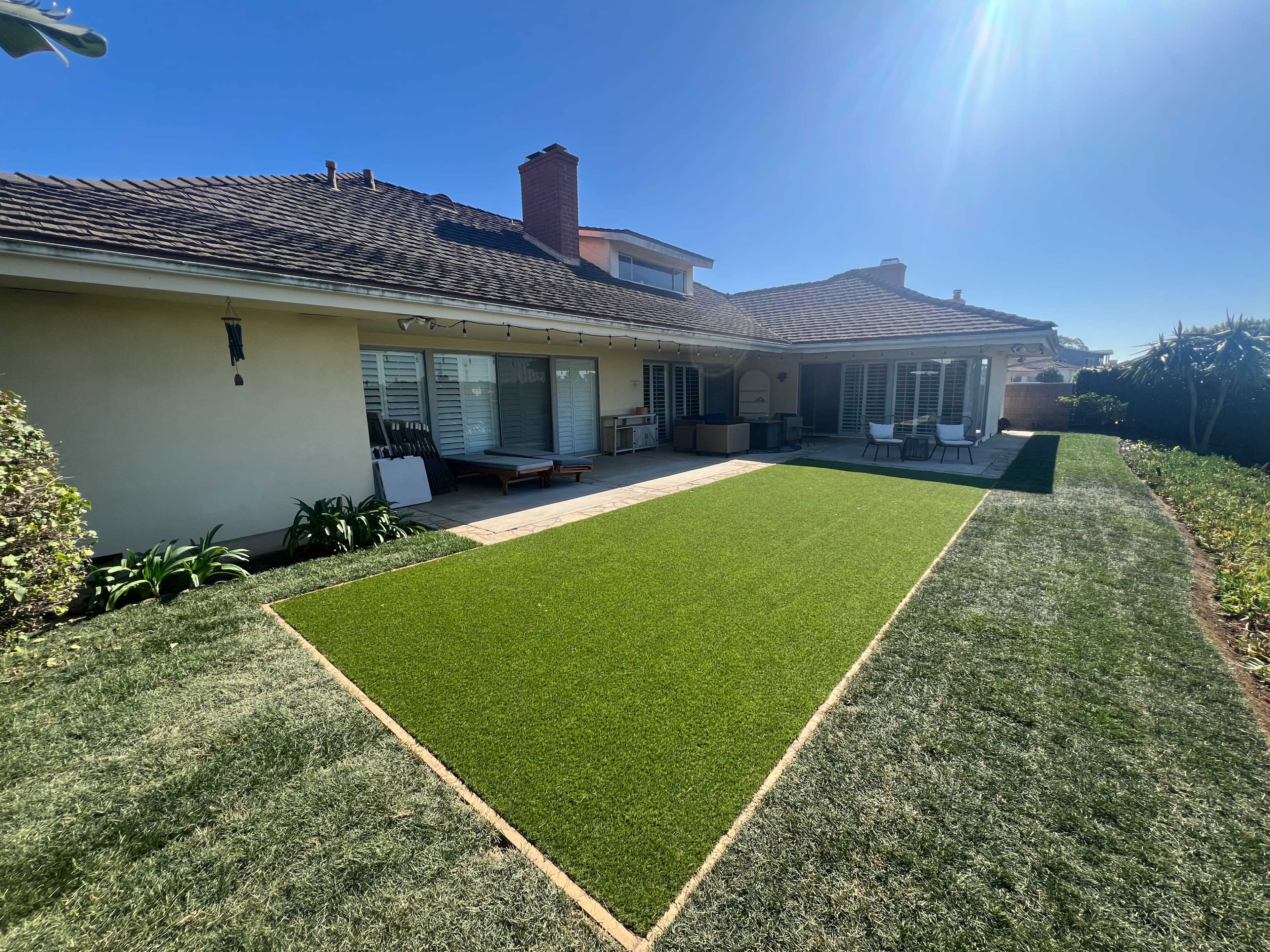 The image shows a well-maintained backyard with artificial grass, a patio area, and a clear blue sky.