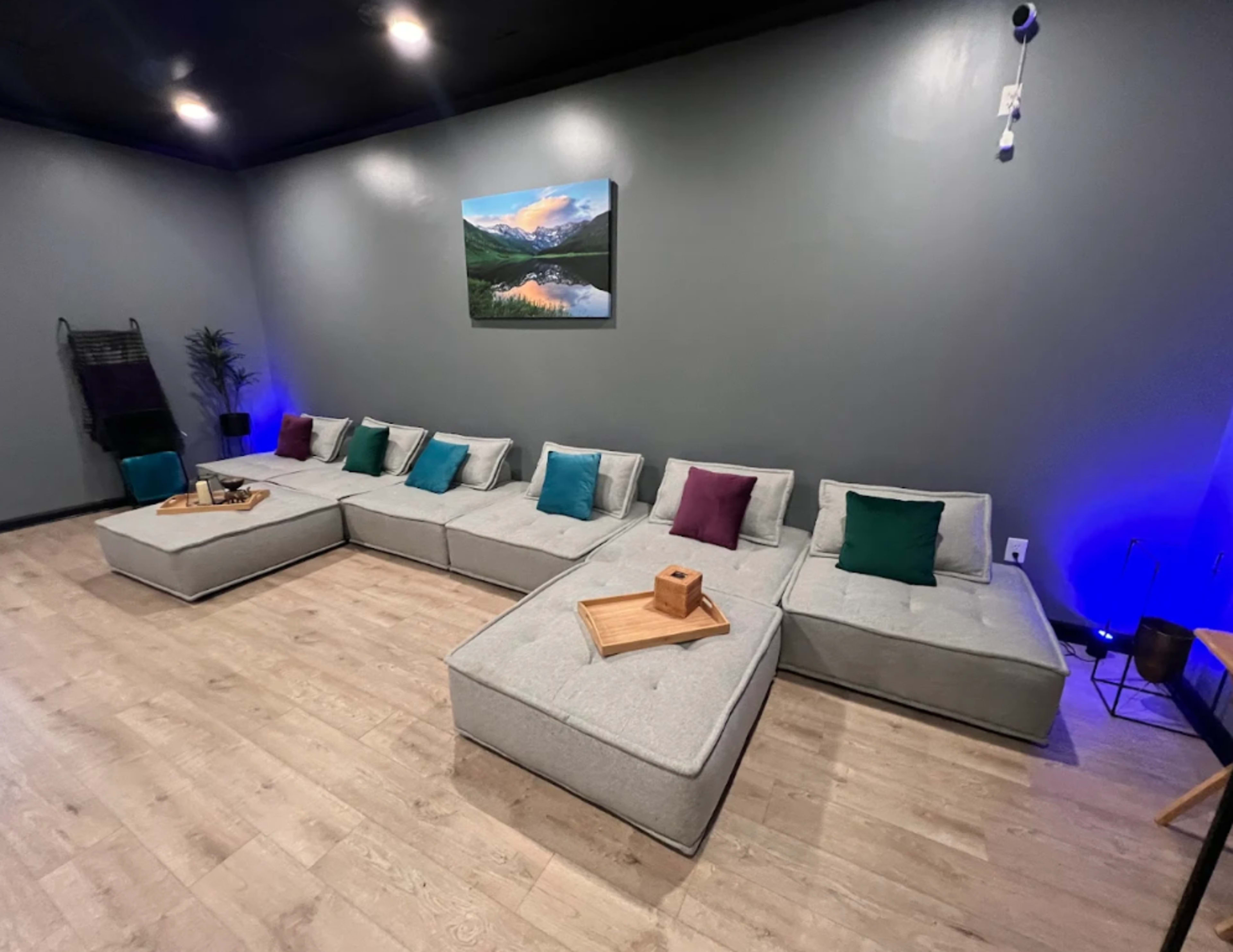 The image shows a modern lounge area featuring a spacious L-shaped sectional sofa with colorful accent cushions and a wooden tray on it, set against a gray wall with a landscape painting.