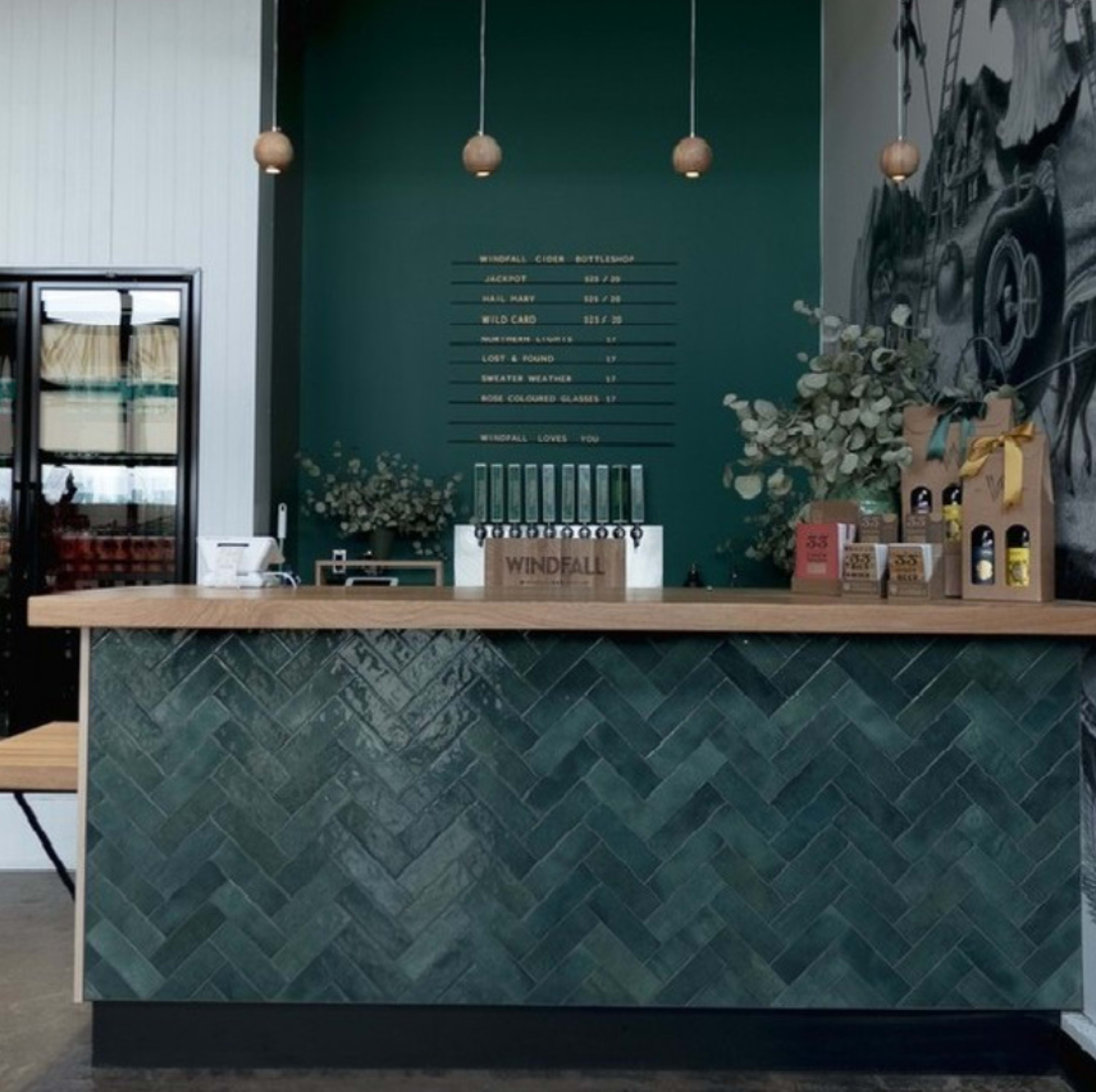 The image shows a modern bar counter with a herringbone-patterned green tile facade, a wooden countertop, and shelves displaying beverages.