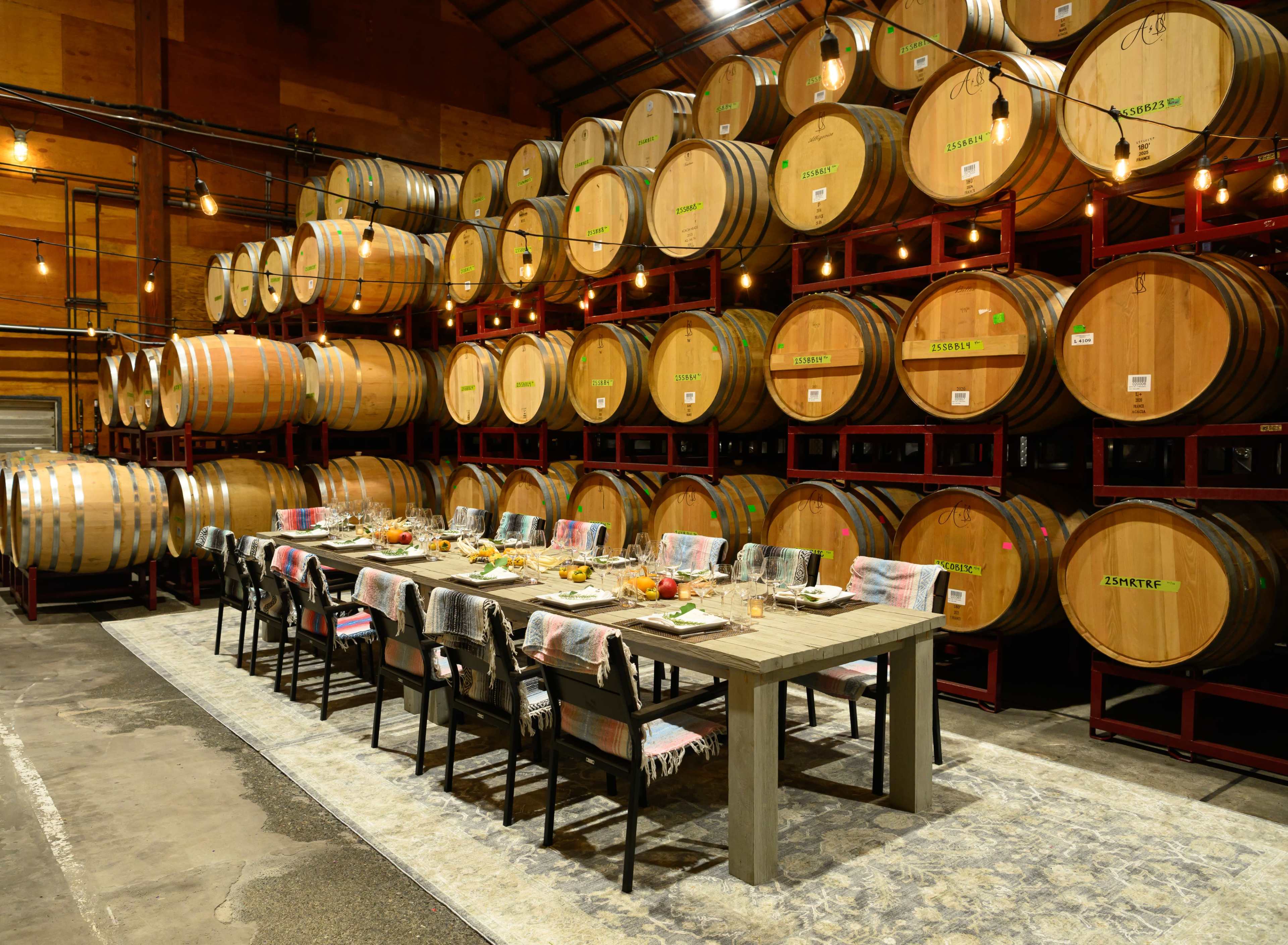 A long dining table is set with dishes and glassware in front of numerous stacked wine barrels in a rustic setting.