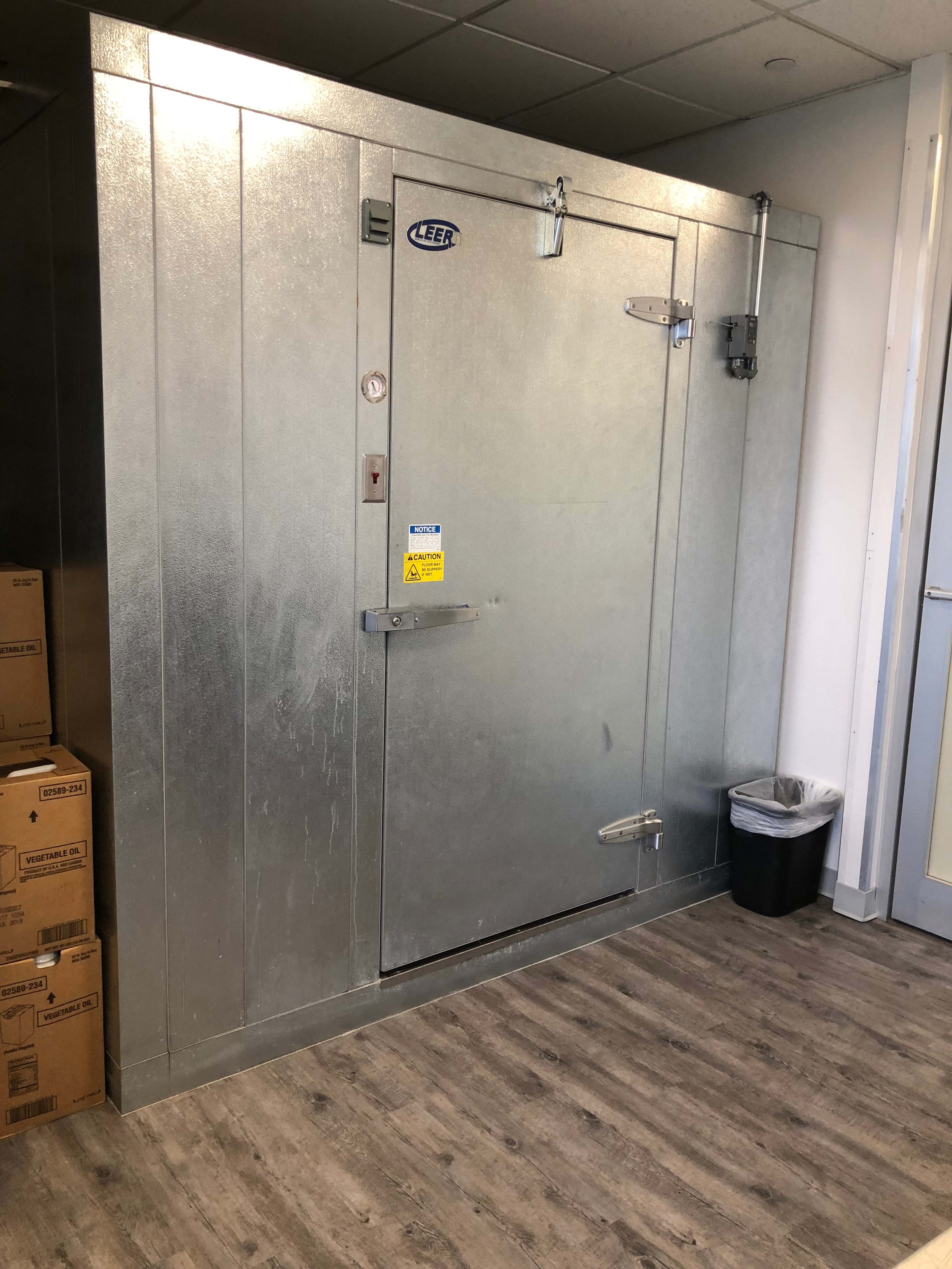 The image shows a metallic walk-in cooler with a large door, located in a room with cardboard boxes stacked nearby and a trash can in the corner.