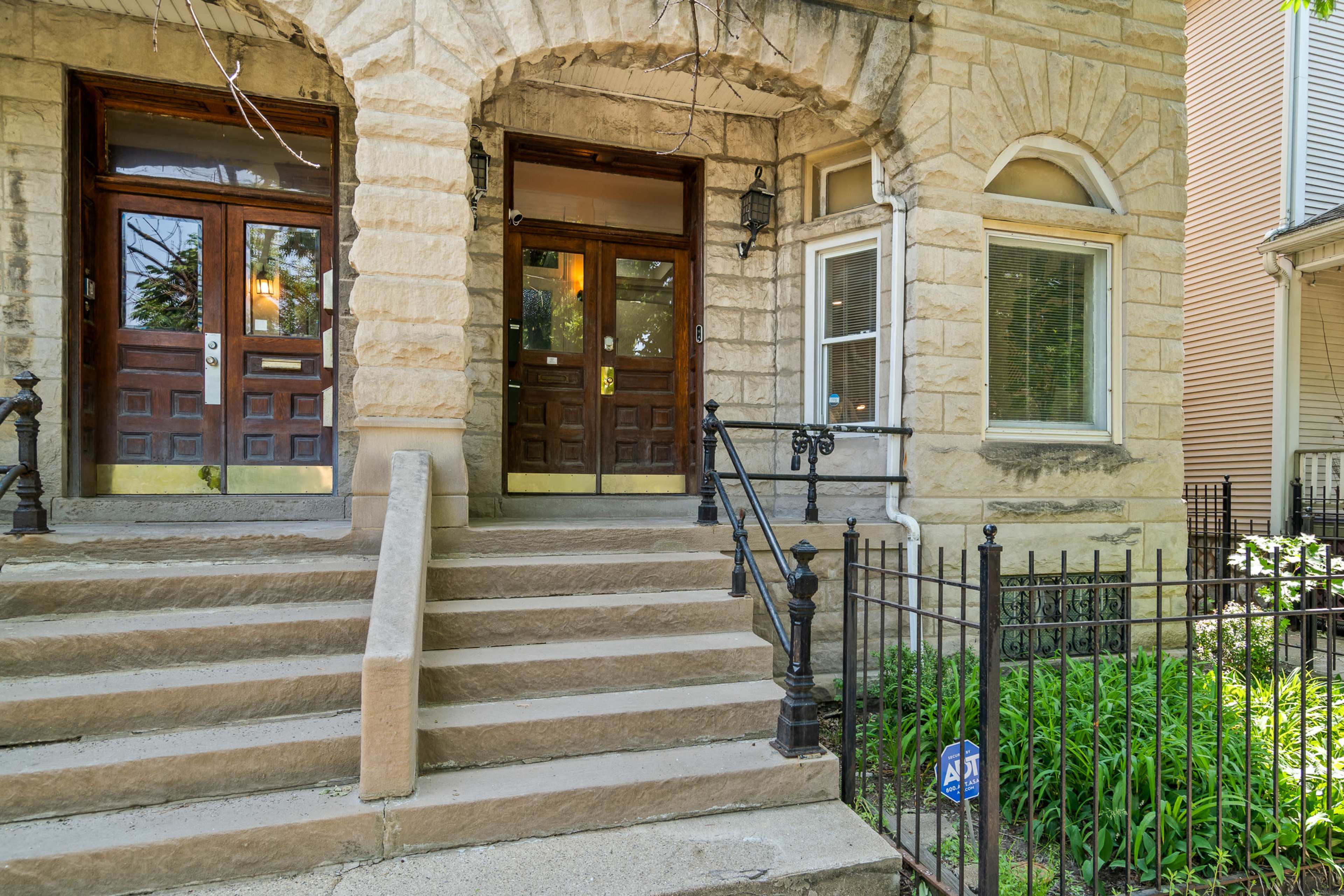 The image shows a stone building entrance with two wooden doors, flanked by steps and a wrought-iron railing, leading to a small garden area.
