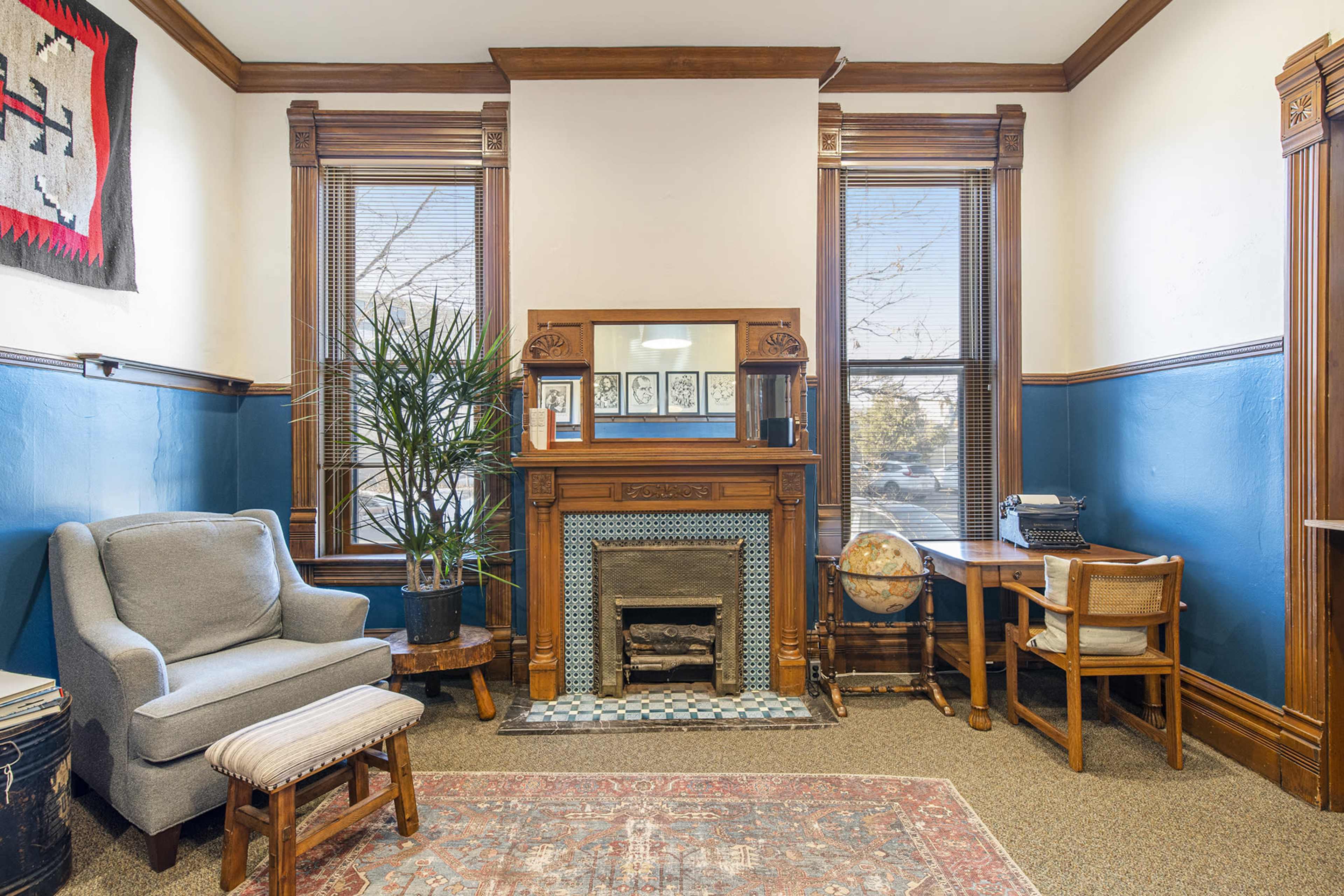 A cozy room featuring a fireplace, a gray armchair, a small wooden table with a globe, and large windows with wooden trim.