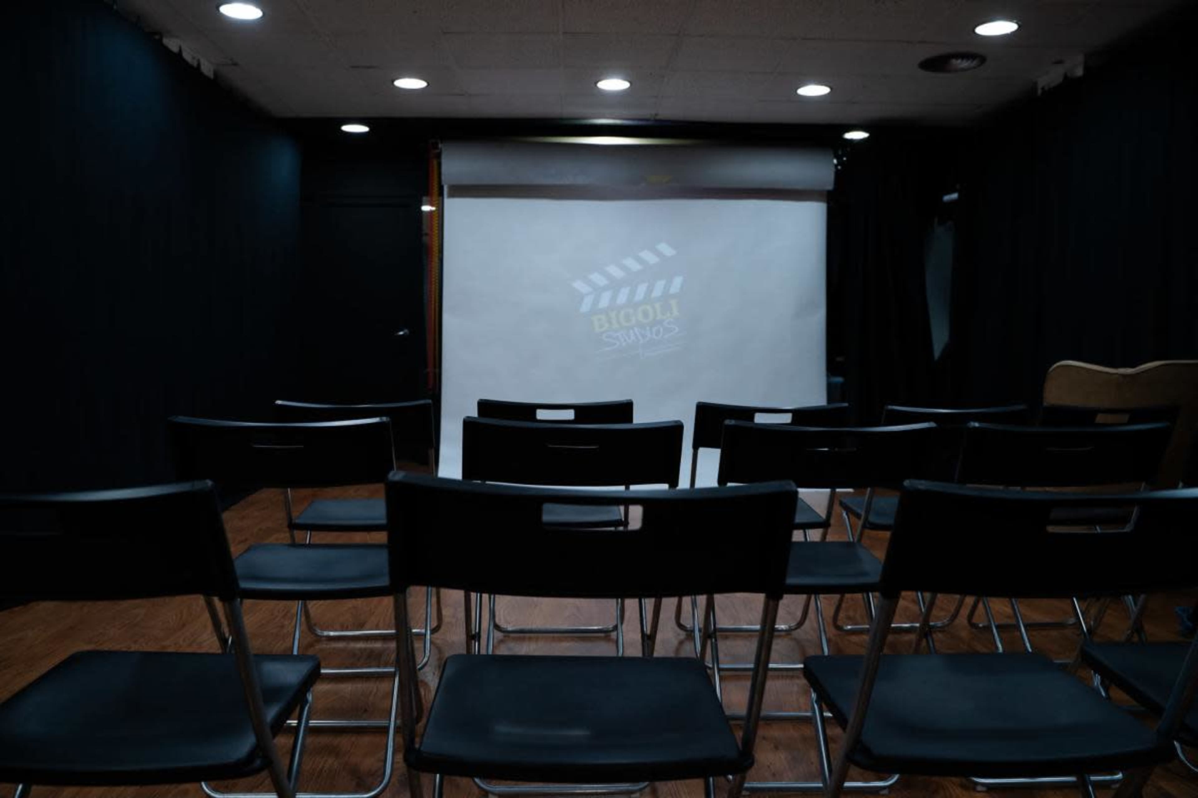 The image shows a row of black folding chairs facing a blank projection screen in a darkened room.
