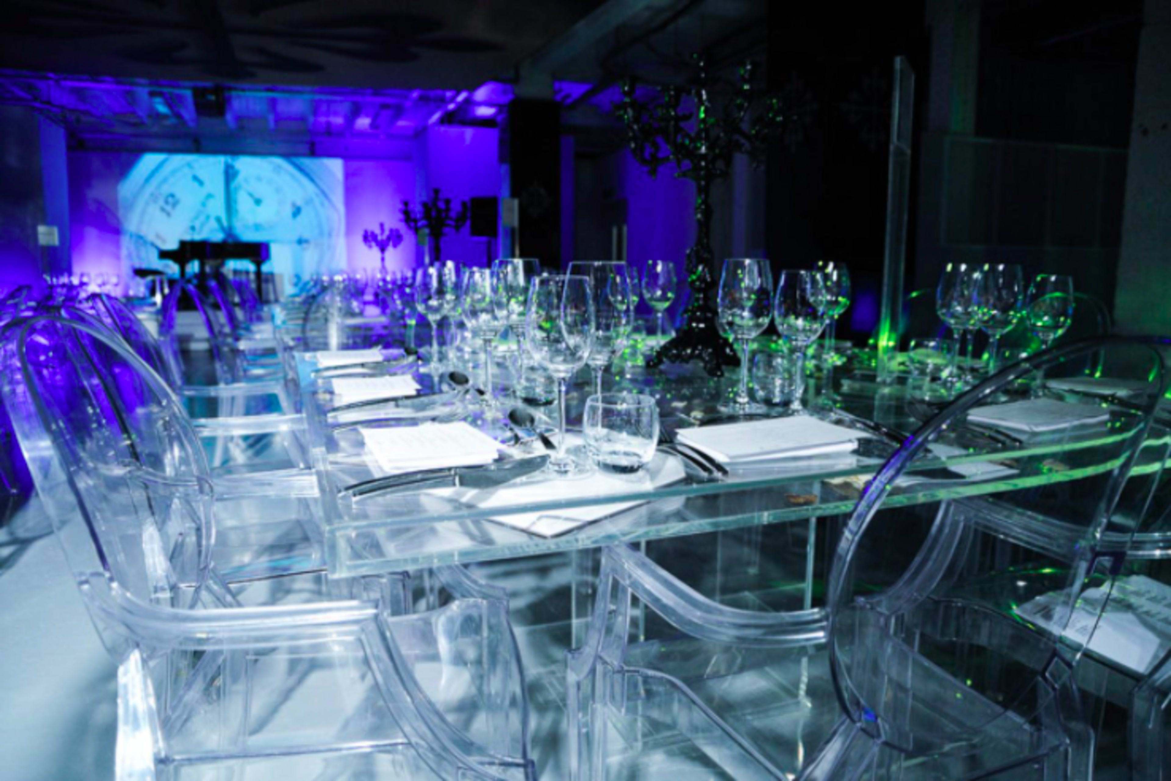 A long dining table is set with clear acrylic chairs and multiple glassware items under purple and green lighting.