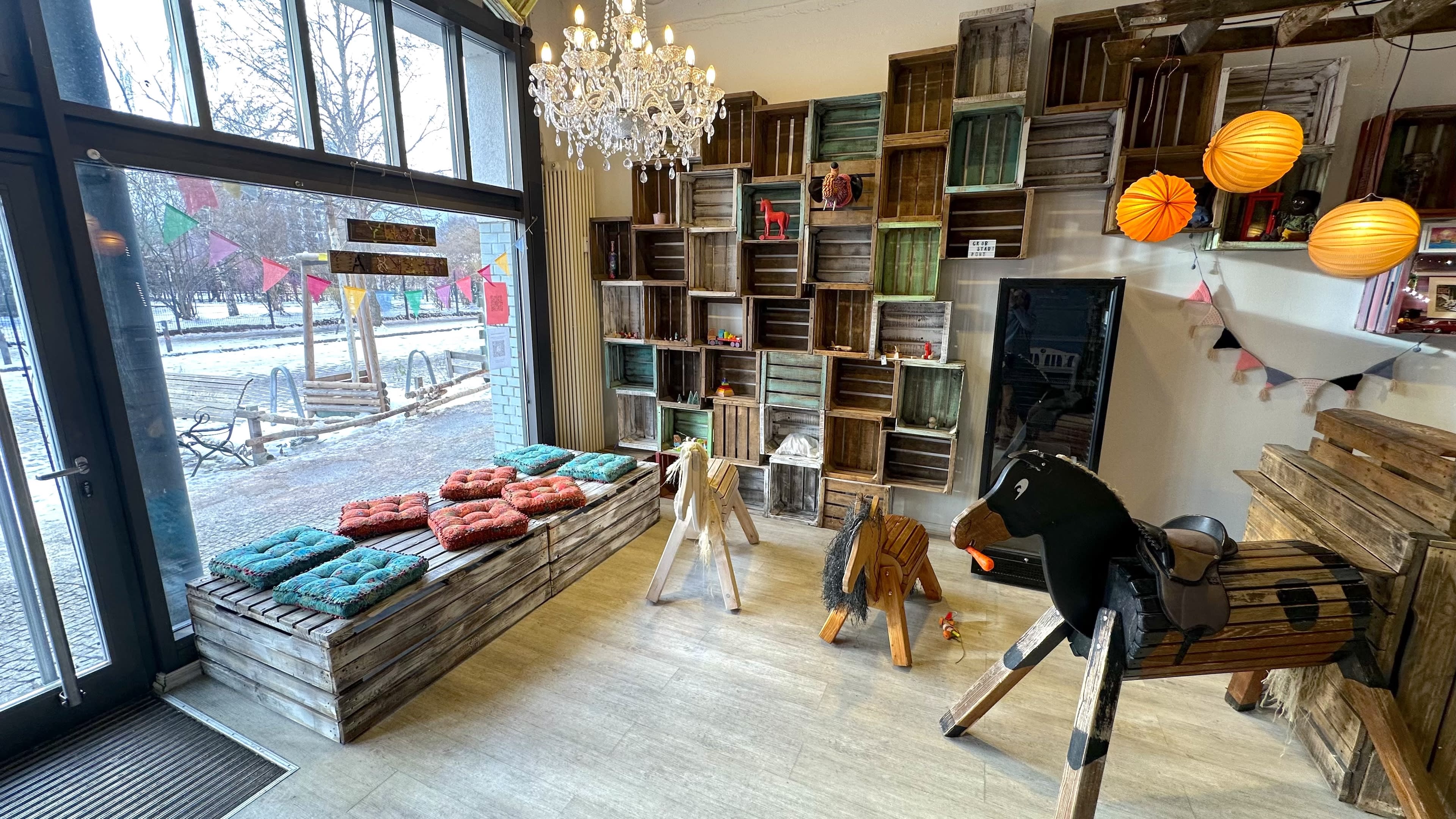 The image shows a cozy interior space with wooden crates used as shelves, colorful cushions on benches, and playful wooden horse sculptures.