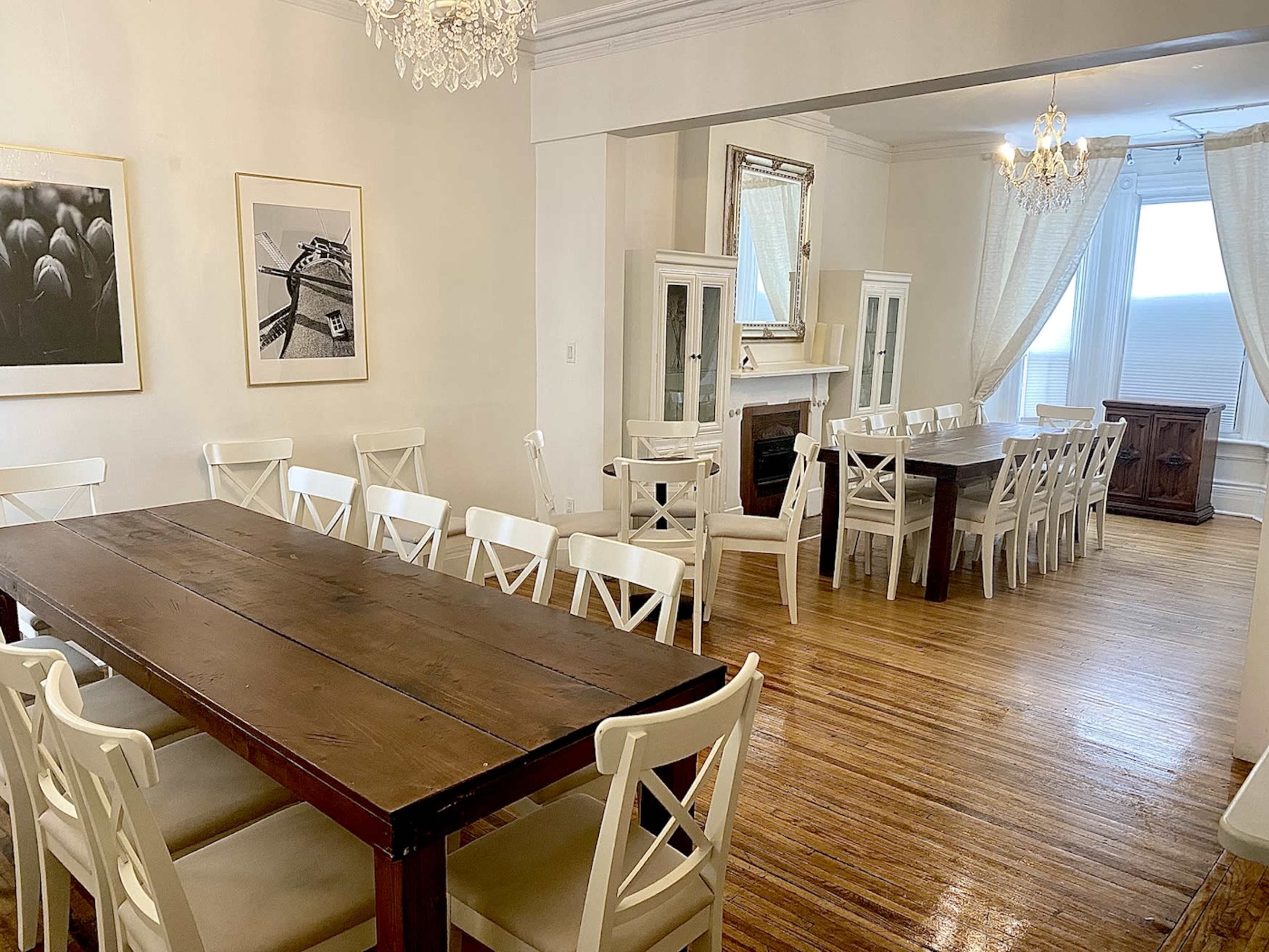 A spacious dining area with two long wooden tables surrounded by white chairs, complemented by large windows and light-colored walls.