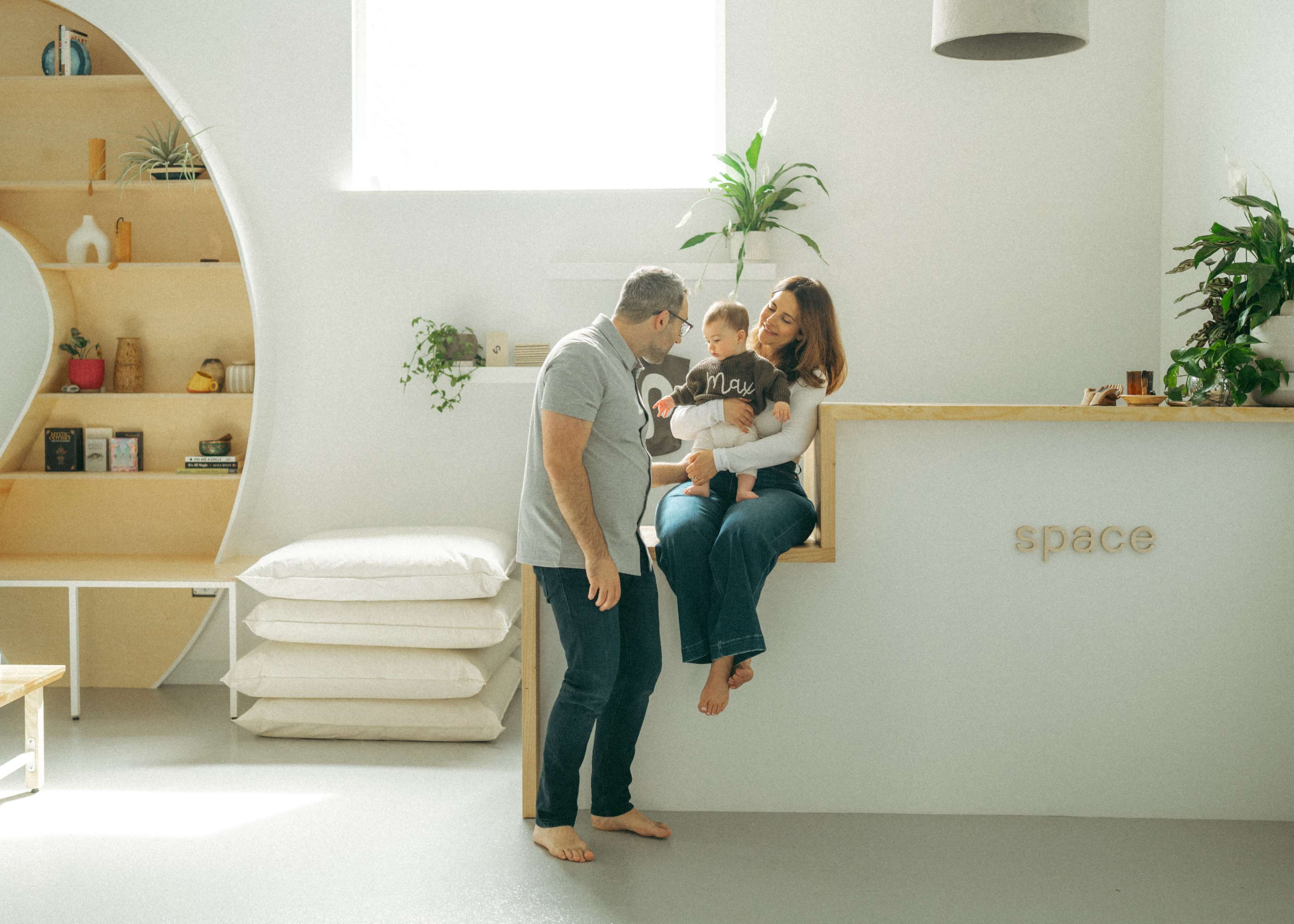A family of three is gathered in a bright, minimalistic living space, with one parent holding a toddler while the other stands nearby.