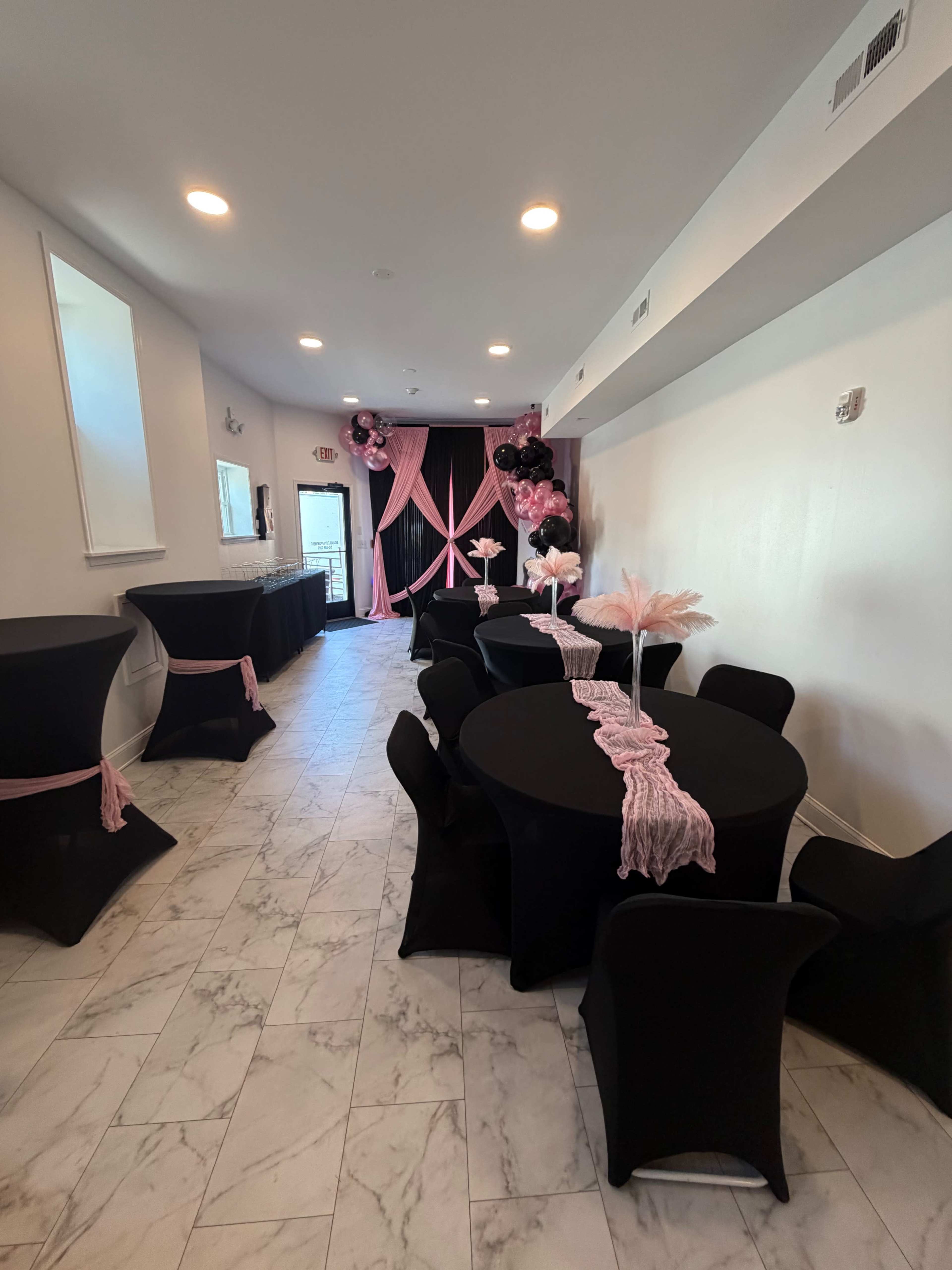 The image shows a decorated event space with black tables and chairs, featuring pink decorations and a backdrop at one end.