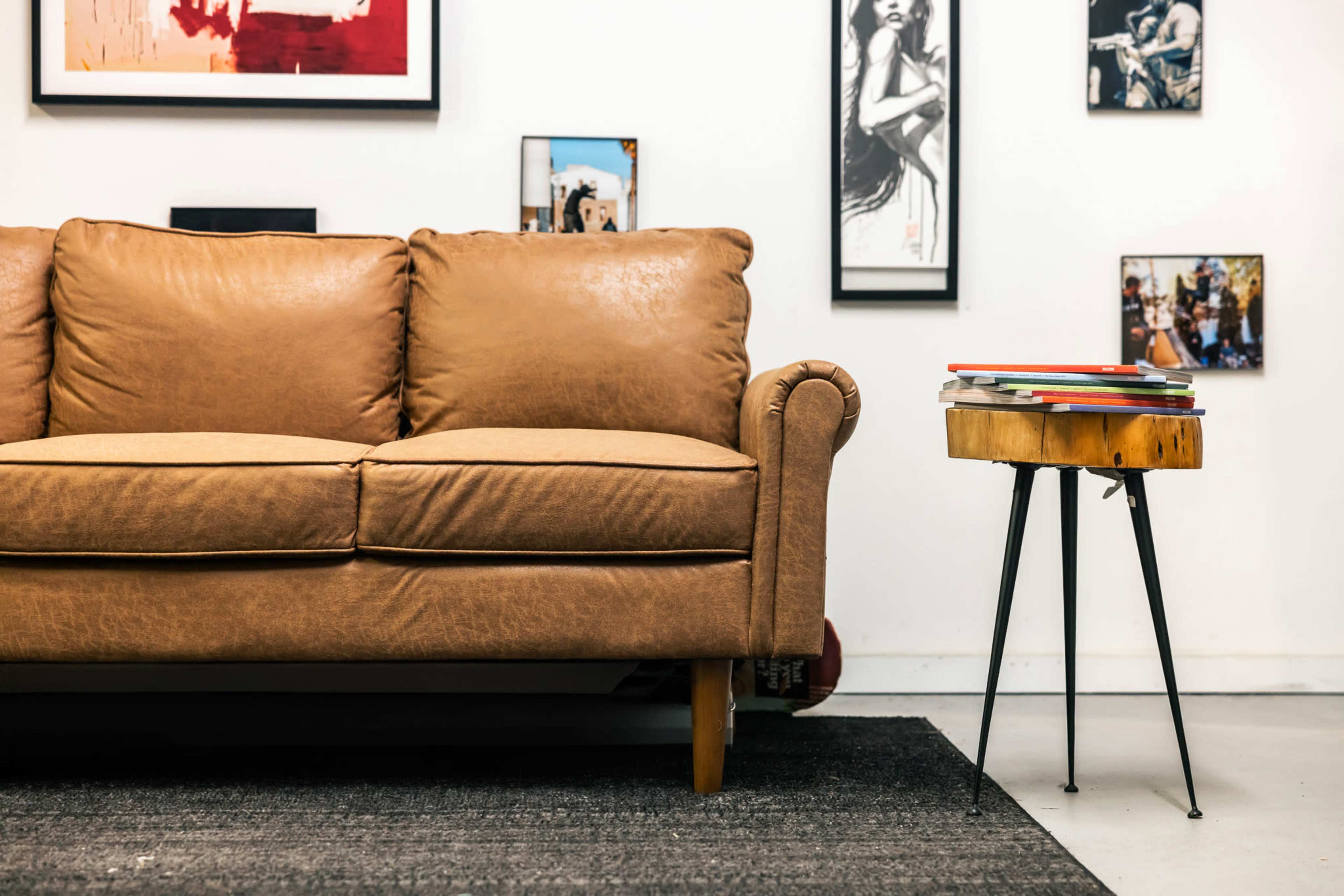 A brown leather couch sits beside a small wooden side table stacked with magazines, against a wall adorned with various framed pictures.