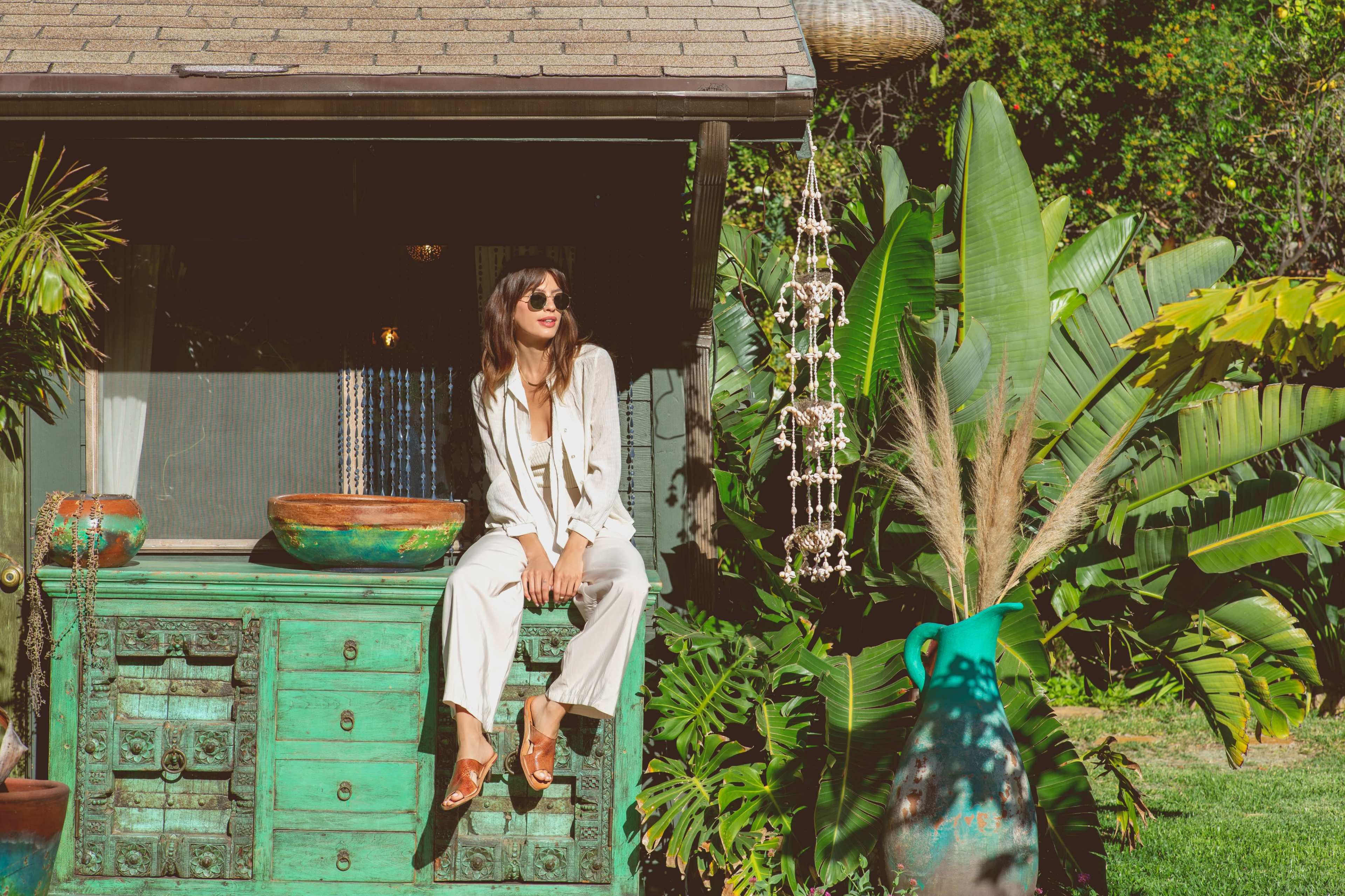 A woman sits on a turquoise cabinet next to tropical plants and decorative items in a sunny outdoor setting.