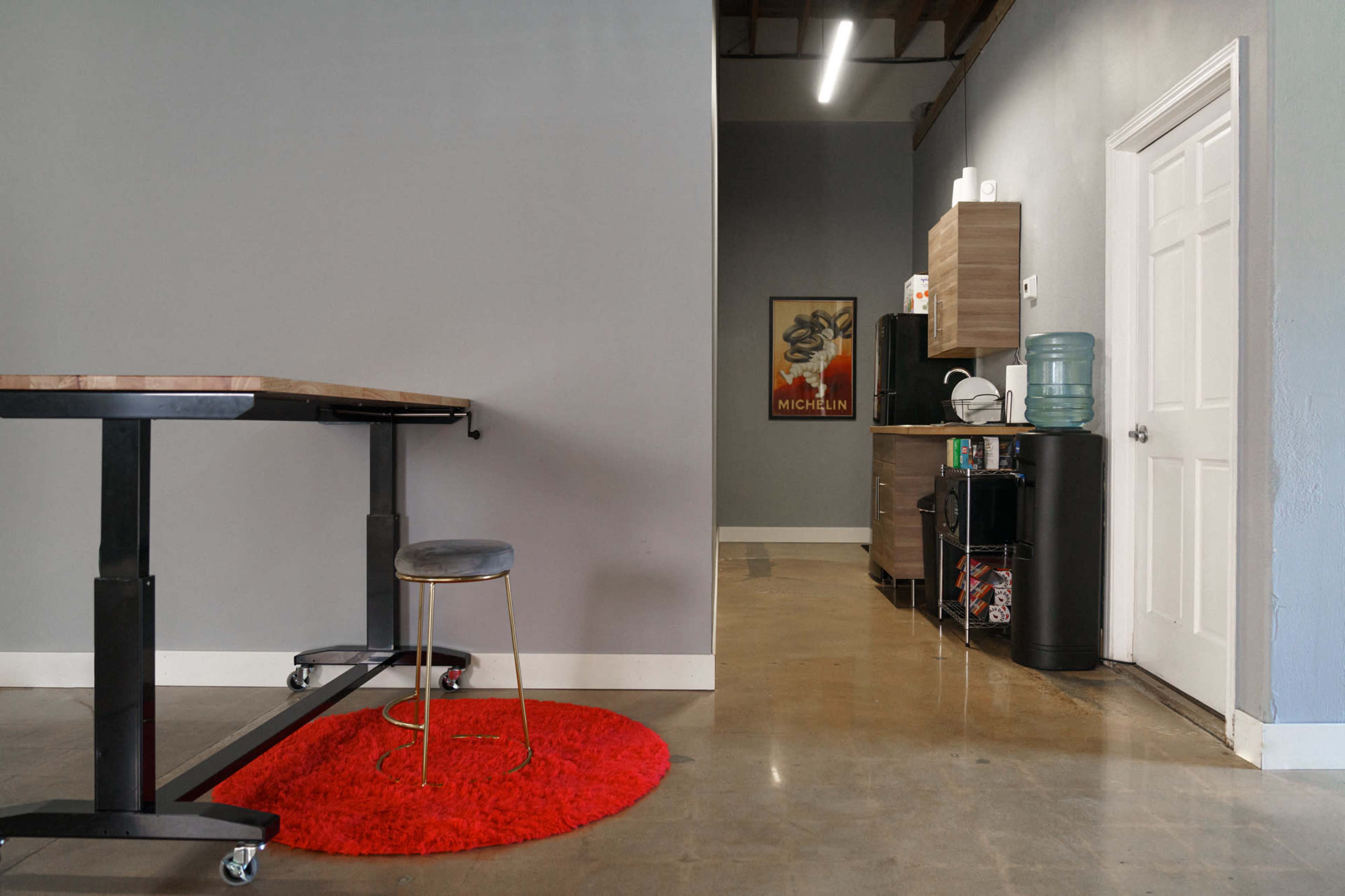 A modern workspace with a wooden table on wheels, a red circular rug, and a kitchenette area featuring a water cooler and cabinets against a gray wall.