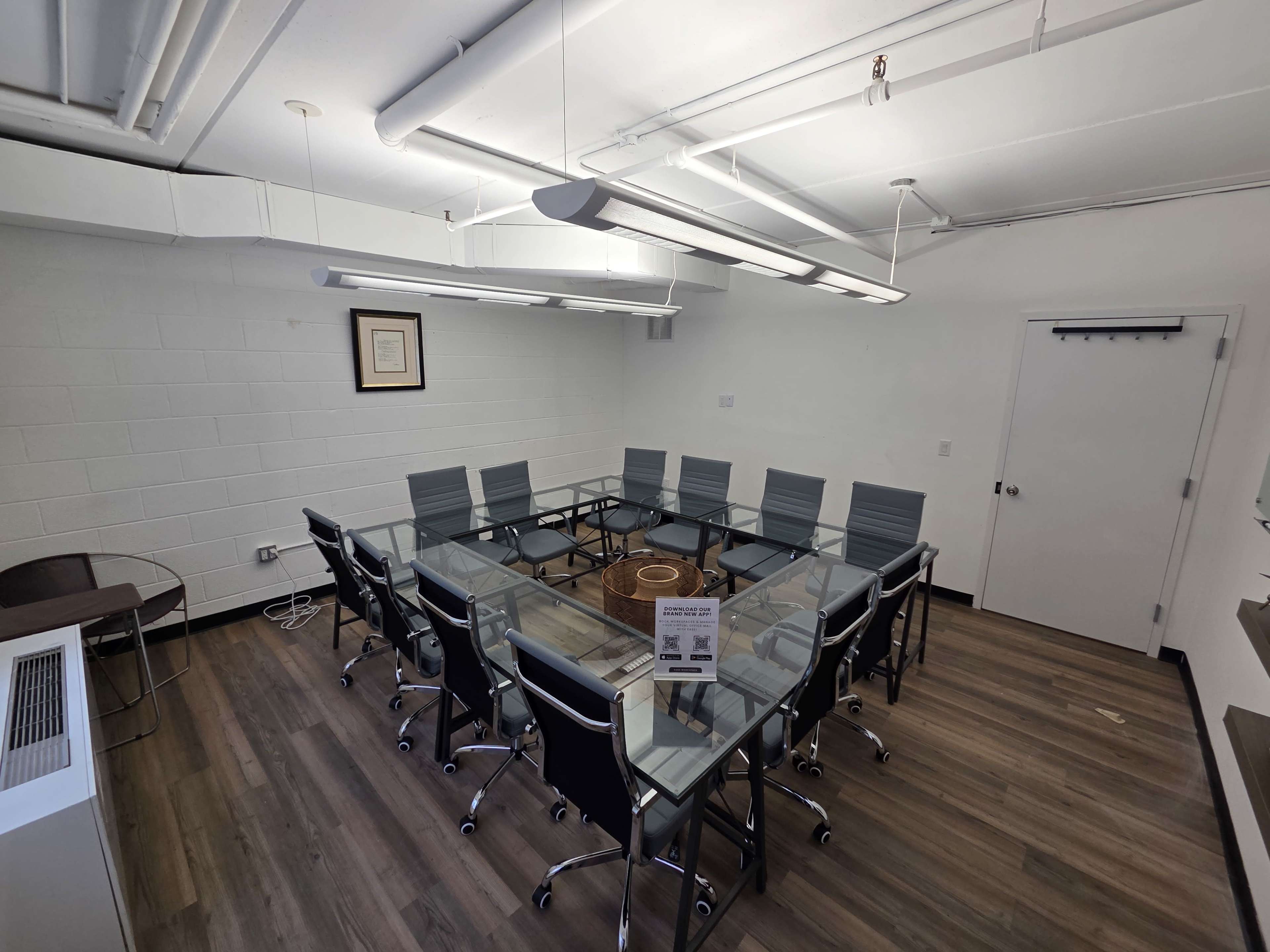 The image shows a modern conference room with a large rectangular glass table surrounded by multiple black and gray chairs, illuminated by overhead lights.