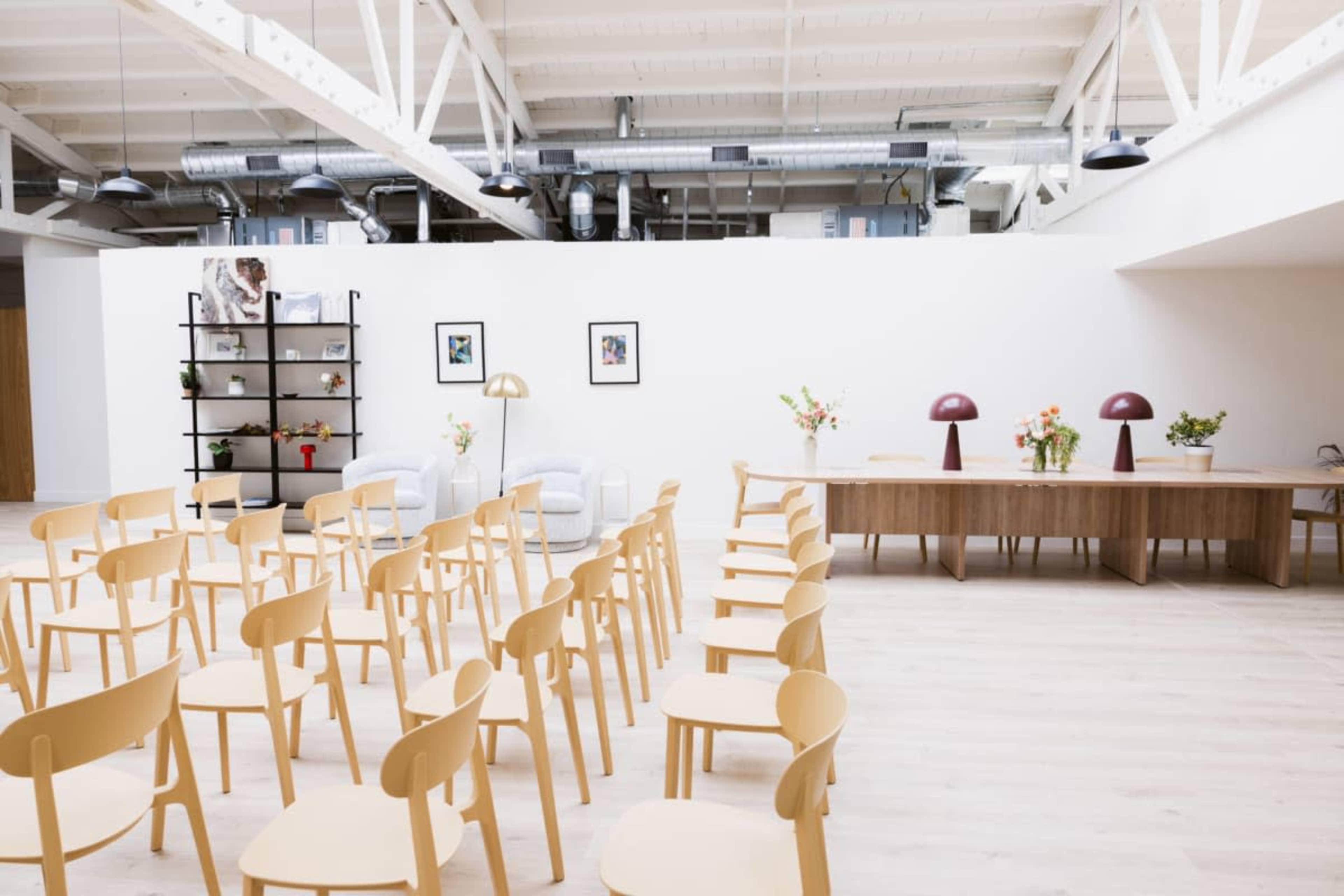 The image shows a bright, spacious room set up for an event, featuring rows of wooden chairs facing a long table and decorative elements like flowers and lamps.
