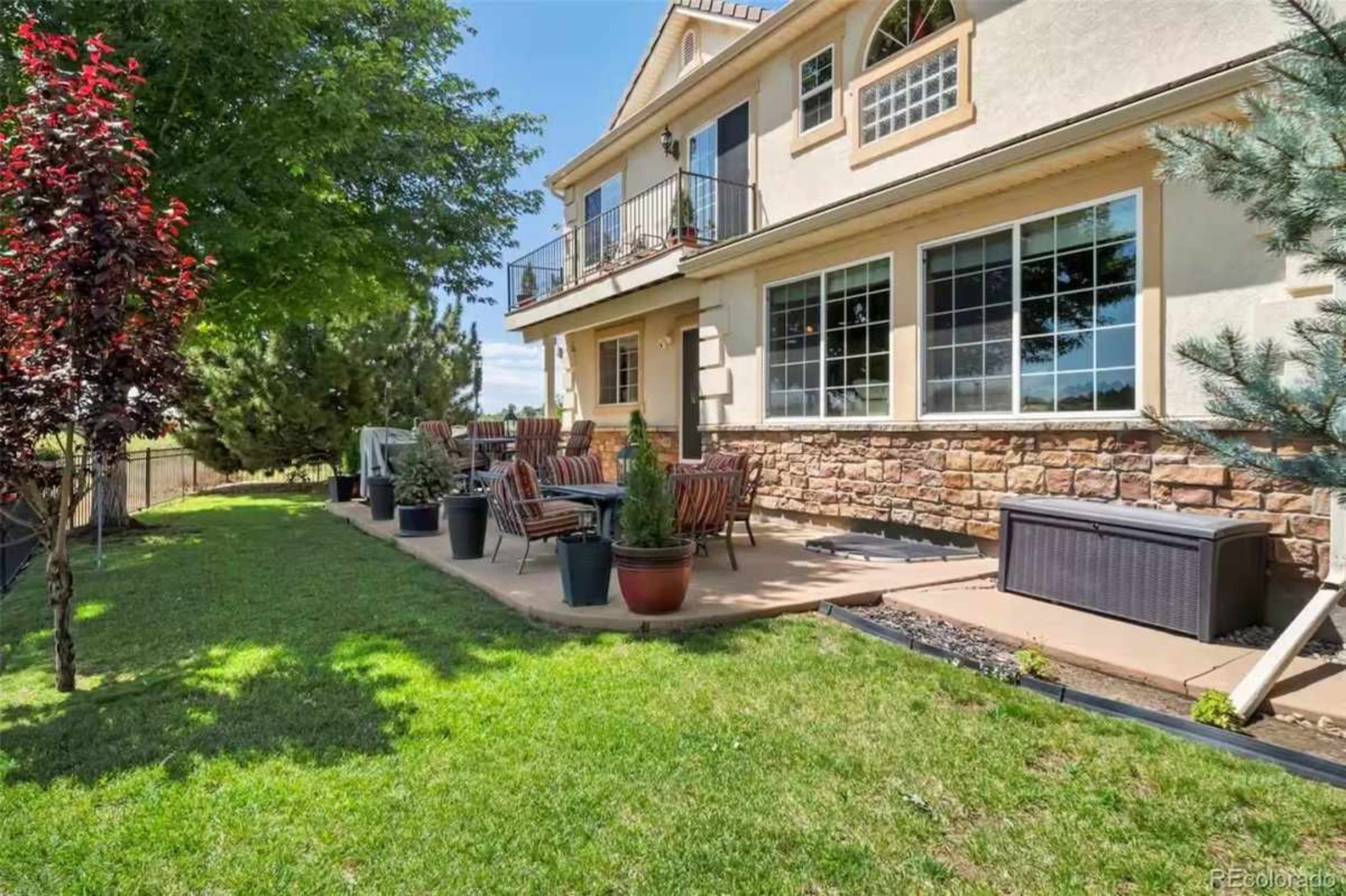 The image shows a spacious backyard patio with outdoor furniture, potted plants, and a stone wall, with a house in the background under a clear blue sky.