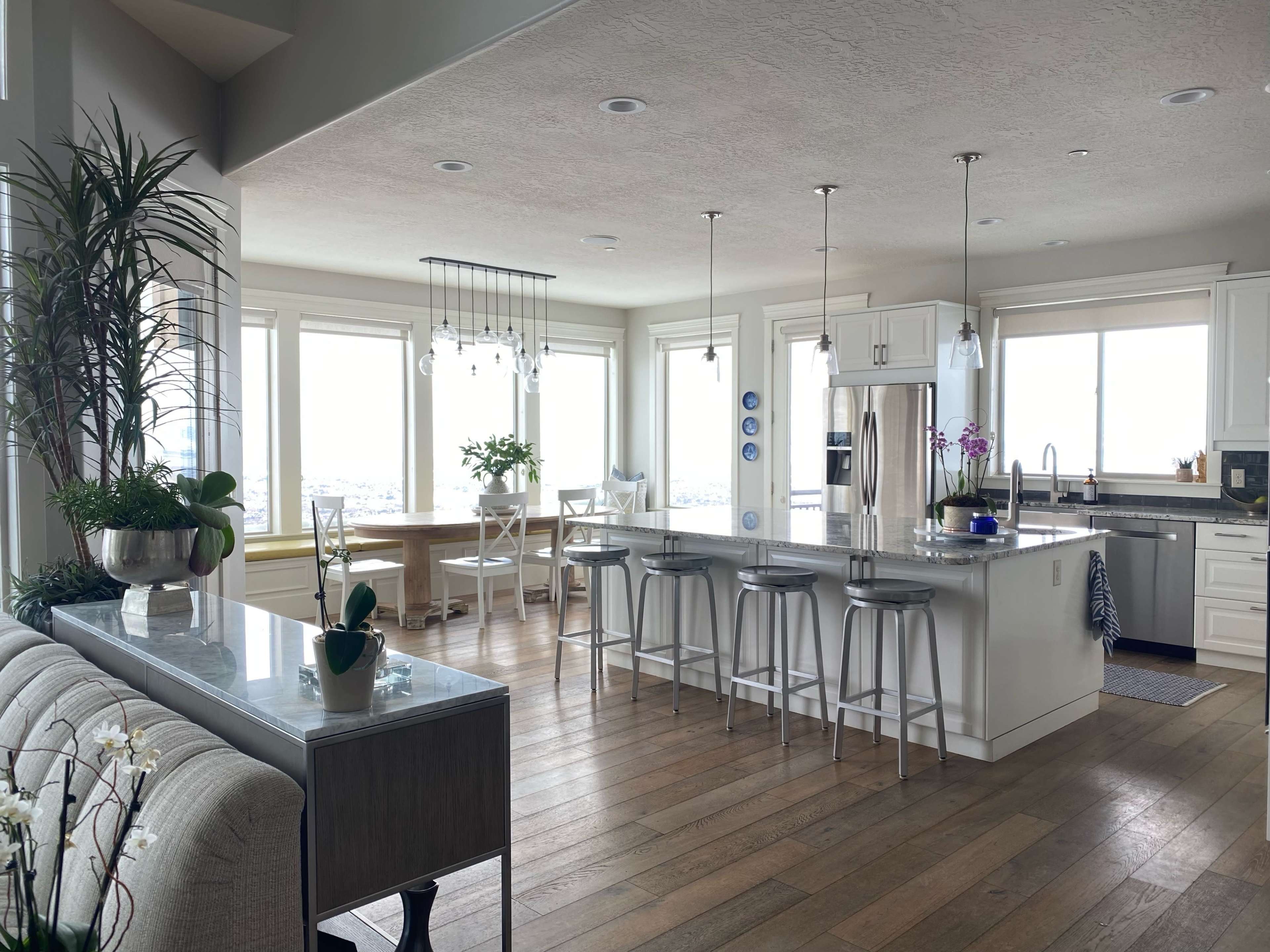 A modern kitchen and dining area feature a large island with bar stools, surrounded by windows that provide a panoramic view of the landscape.