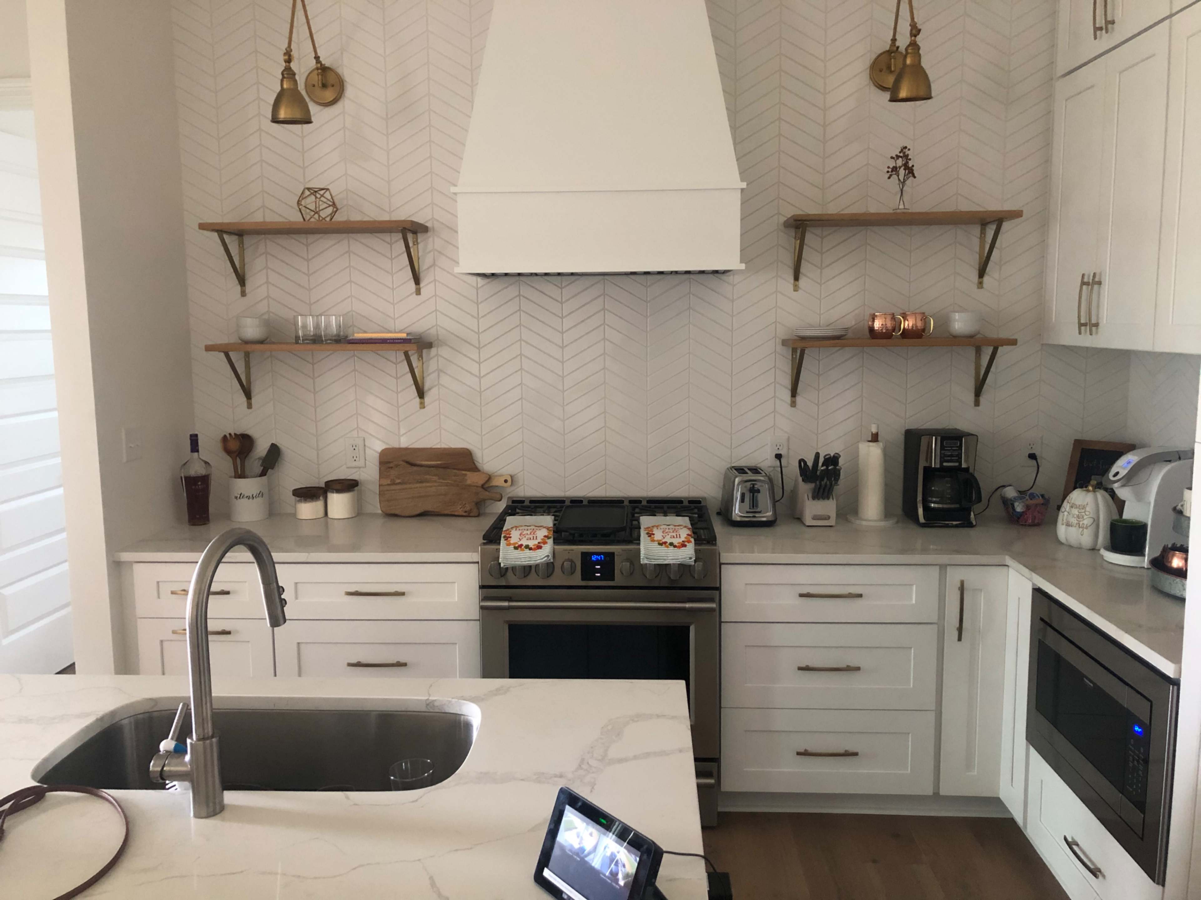 A modern kitchen features a central island with a sink, stainless steel appliances, and herringbone-patterned wallpaper, along with open shelving displaying various kitchen items.