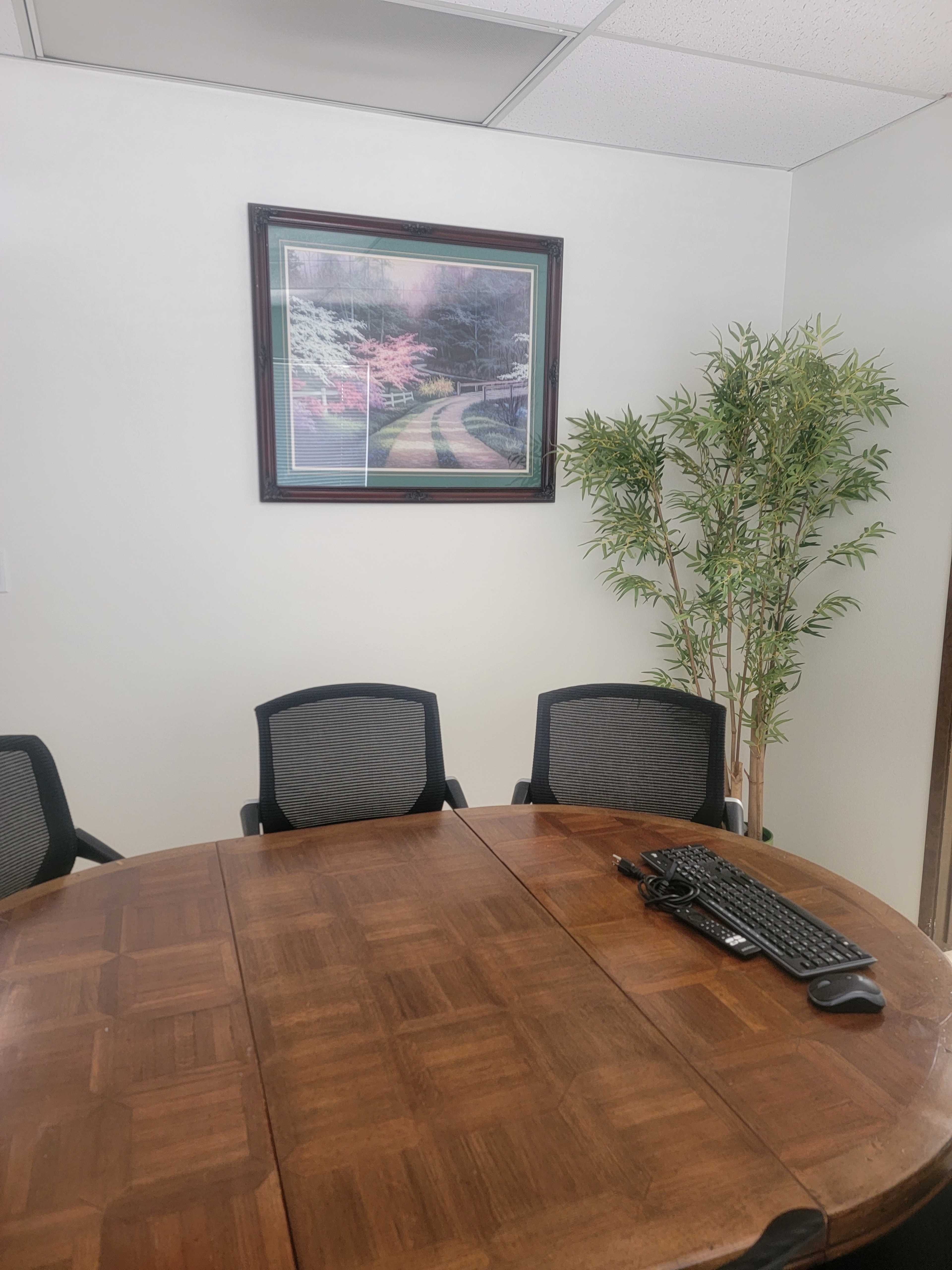A conference room with a circular wooden table, three black chairs, a laptop keyboard, a potted plant, and a framed picture on the wall.