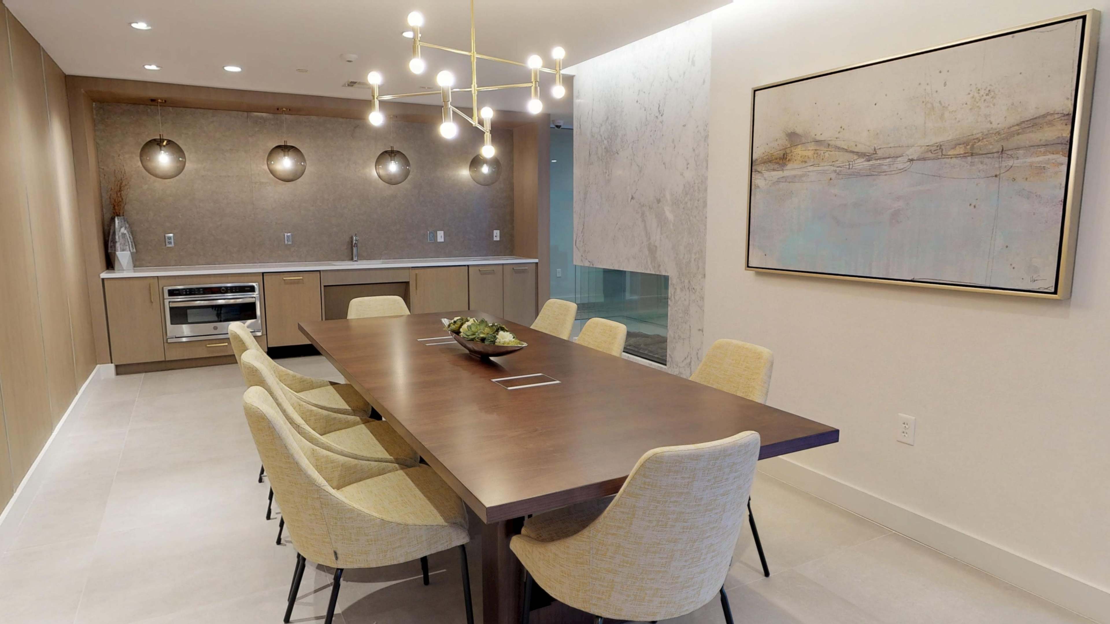 A modern dining area features a long wooden table surrounded by yellow upholstered chairs, with a marble accent wall and contemporary lighting fixtures above.