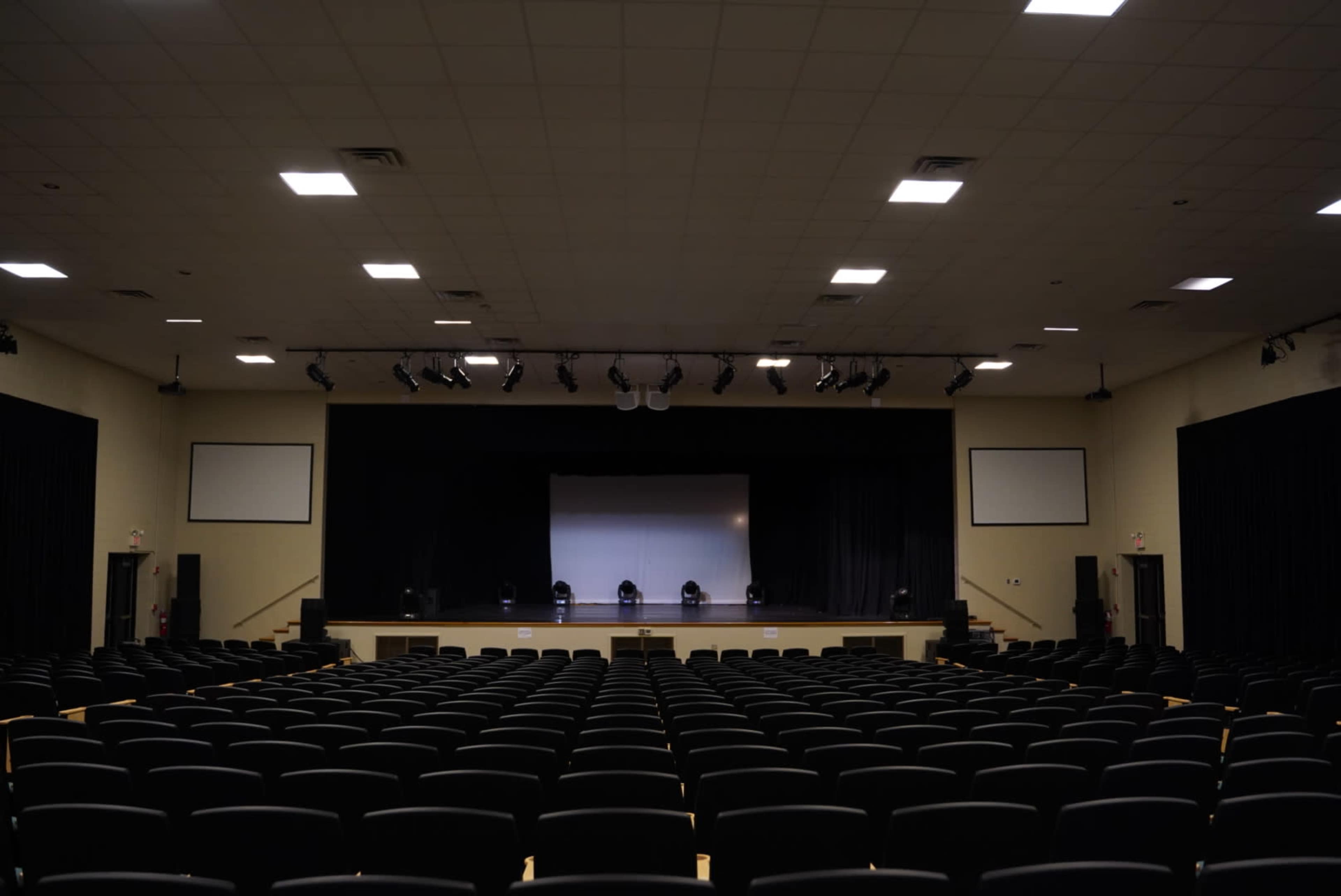 A large auditorium with multiple rows of black seats facing a stage that has a central screen and spotlights.