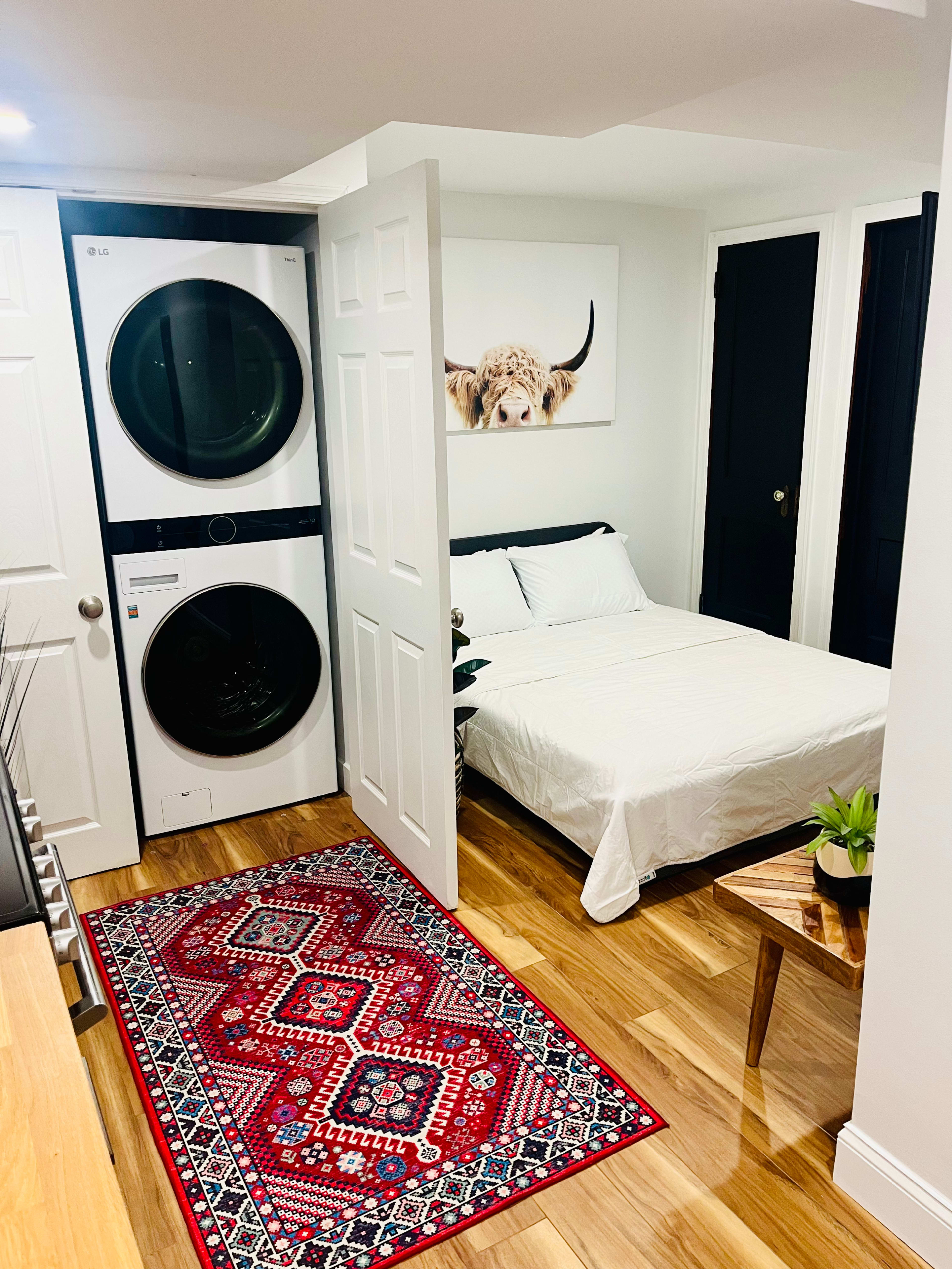 The image shows a small room that features a stackable washer and dryer, a bed with white bedding, a red patterned rug, and a decorative wall art of a cow's head.