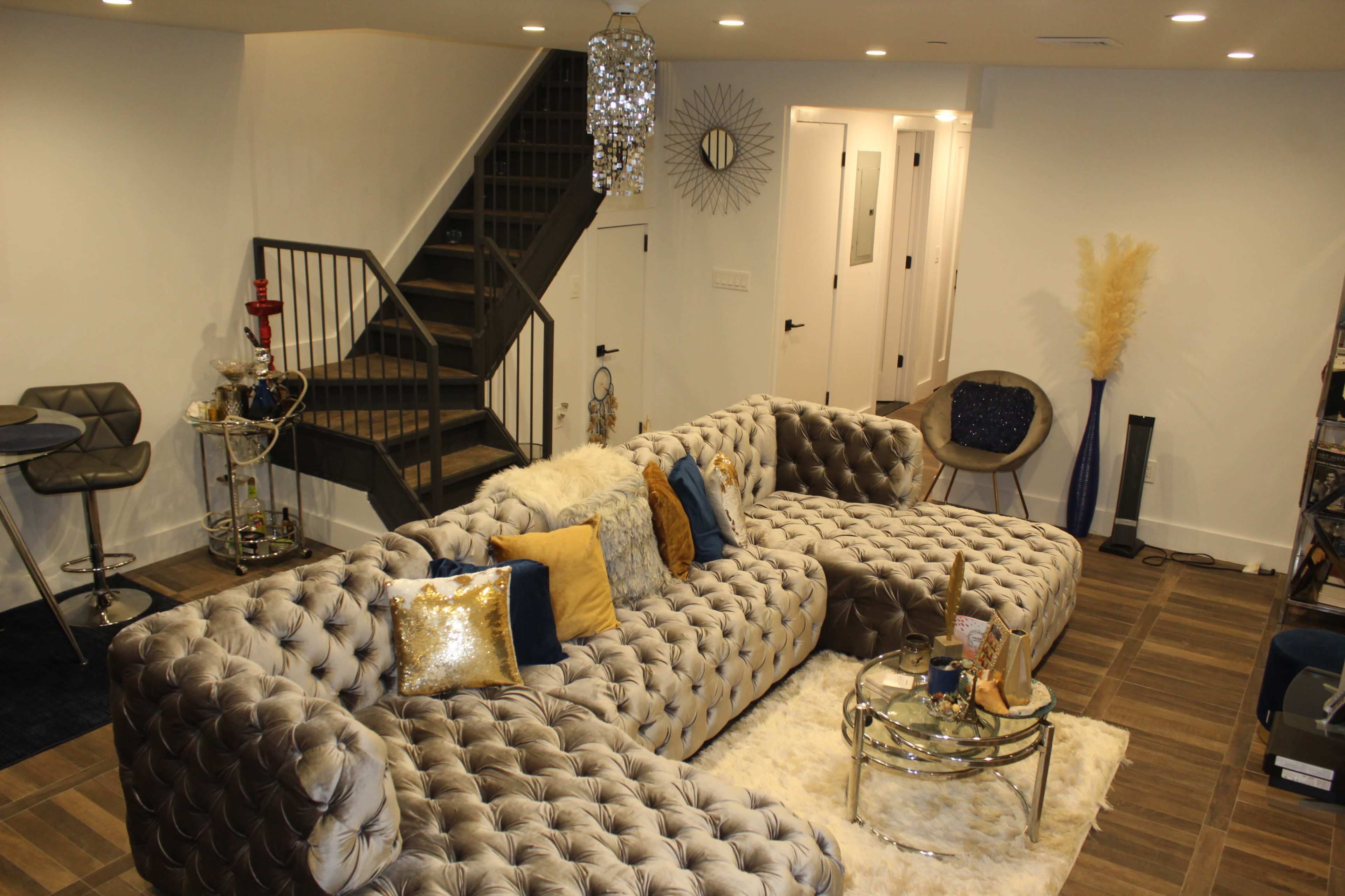 A modern living room featuring a tufted gray sectional sofa, a glass coffee table, and a staircase in the background.