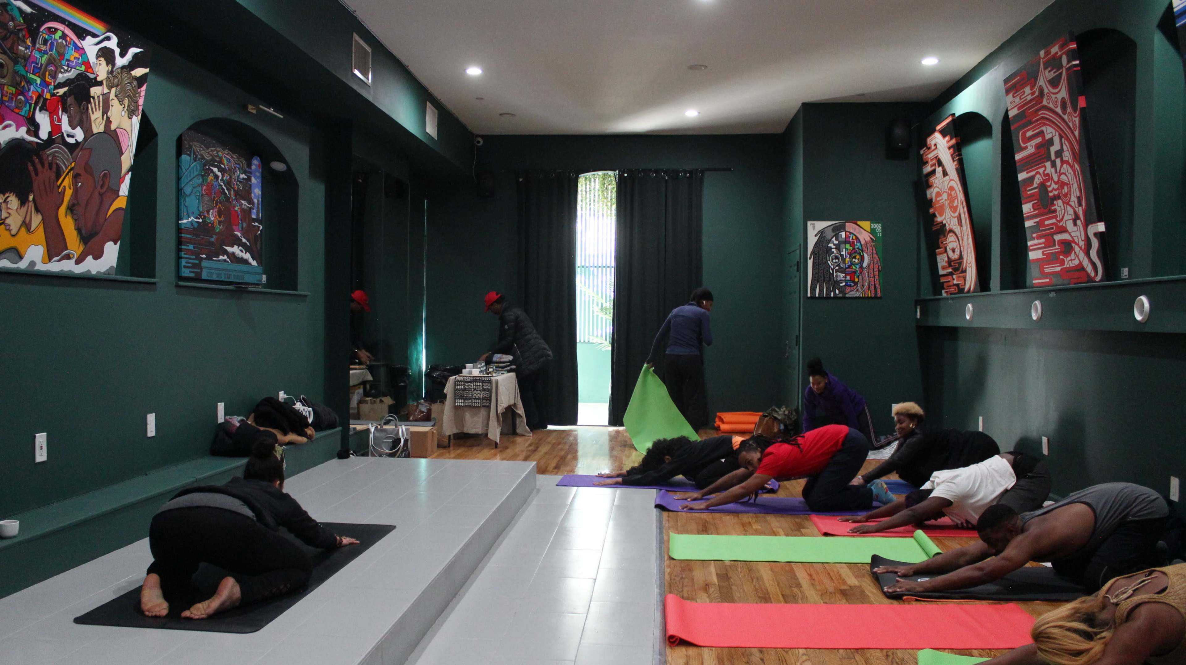 A group of people participates in a yoga session inside a studio featuring dark green walls and colorful artwork.