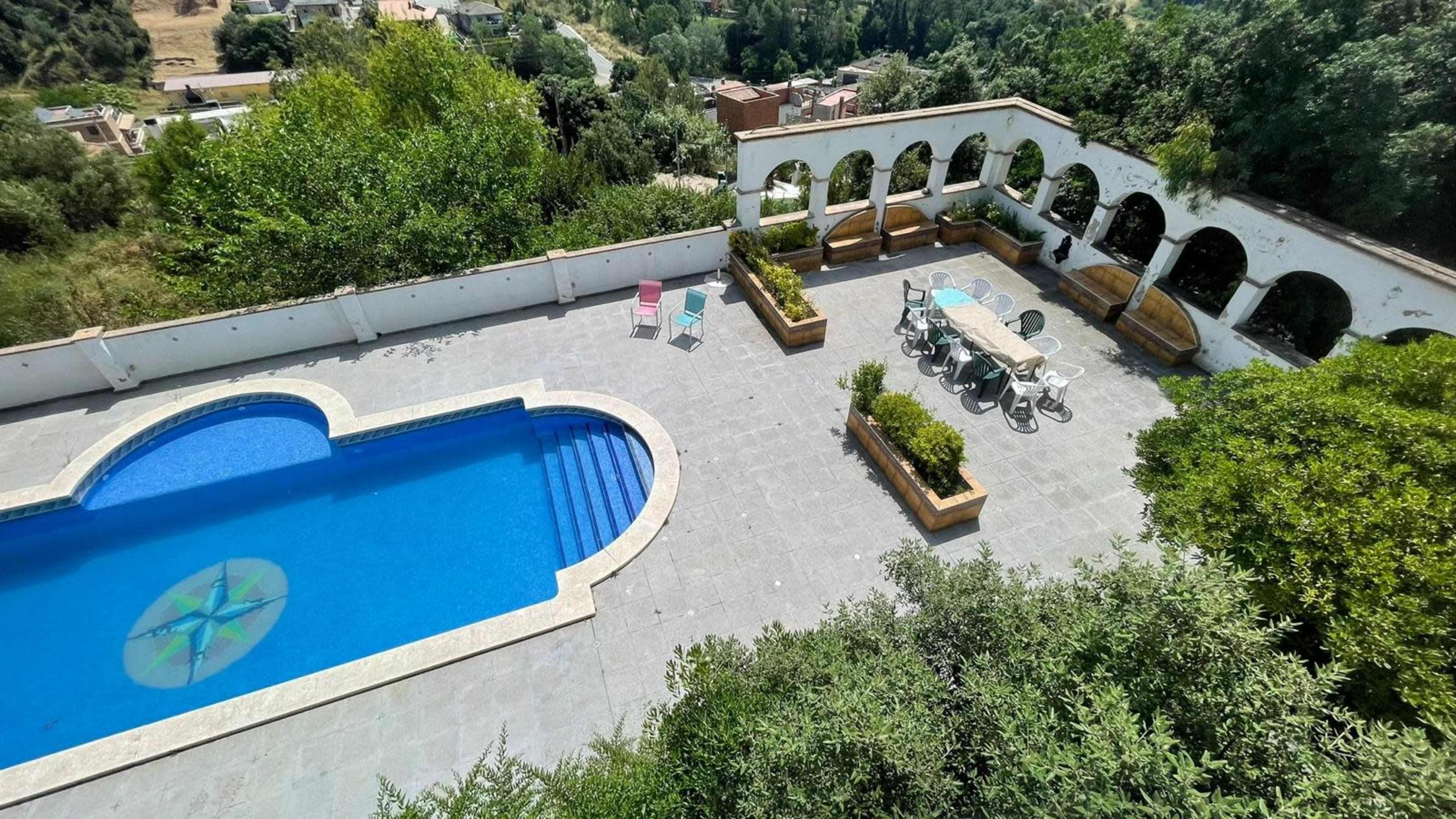 The image shows a spacious outdoor area featuring a swimming pool, a dining table, and garden seating surrounded by greenery.