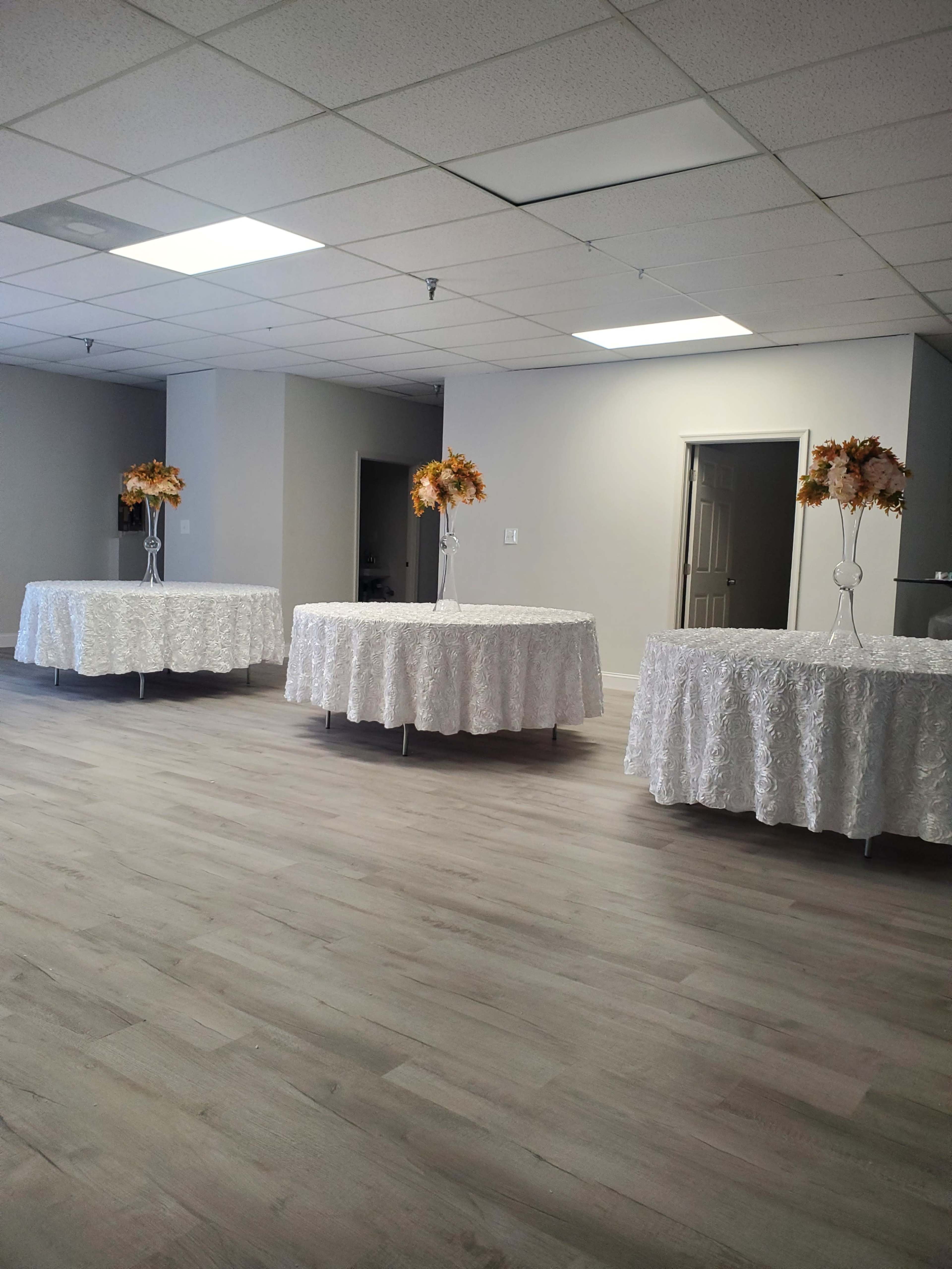 The image shows a spacious interior with four round tables covered in white tablecloths, each topped with a floral arrangement in clear vases.