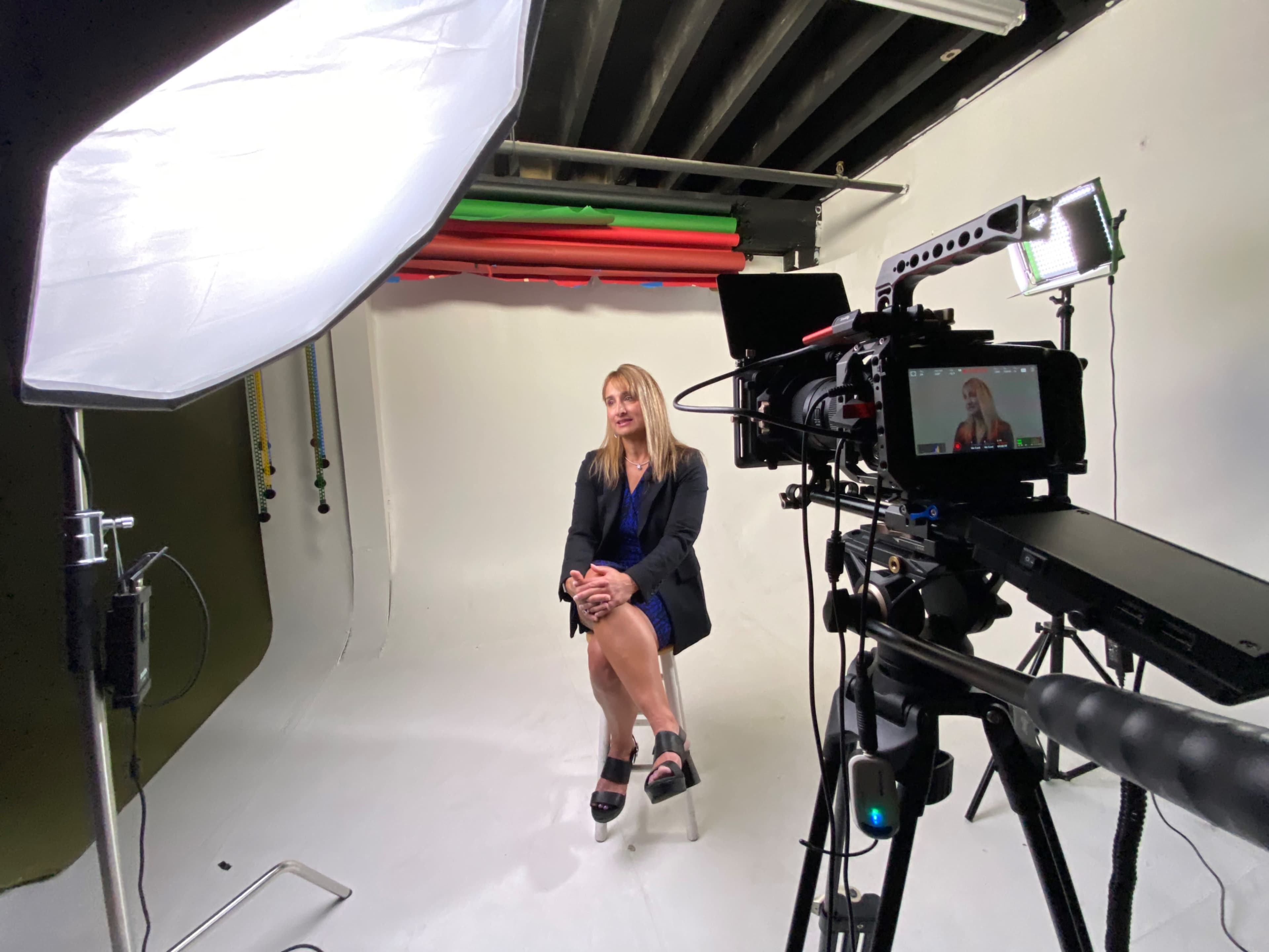 A woman sits on a chair in a studio setup, surrounded by lighting equipment and a camera.