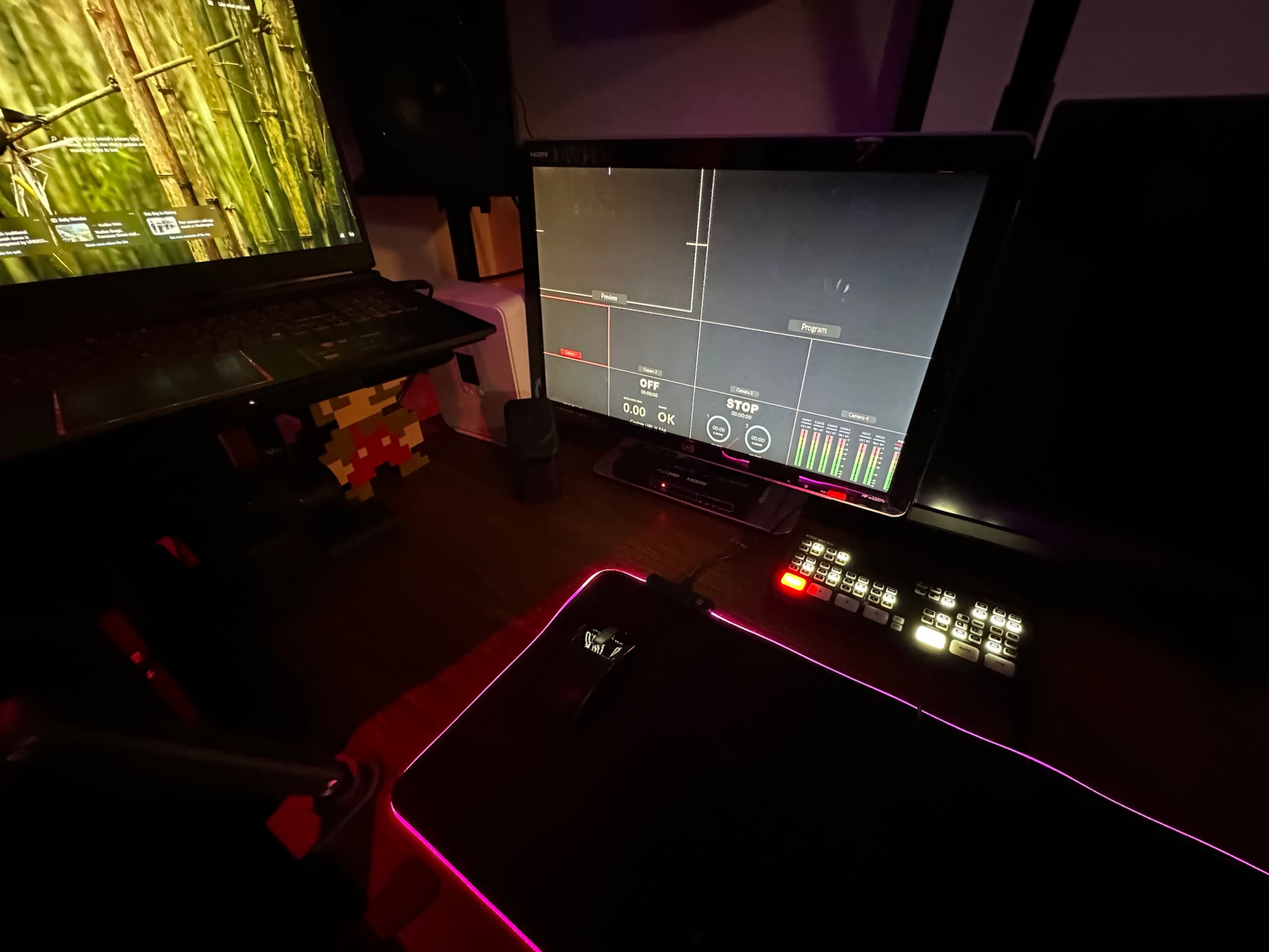 A gaming setup features a monitor displaying audio levels, a laptop, a keyboard with lights, and a mouse on a glowing mousepad.