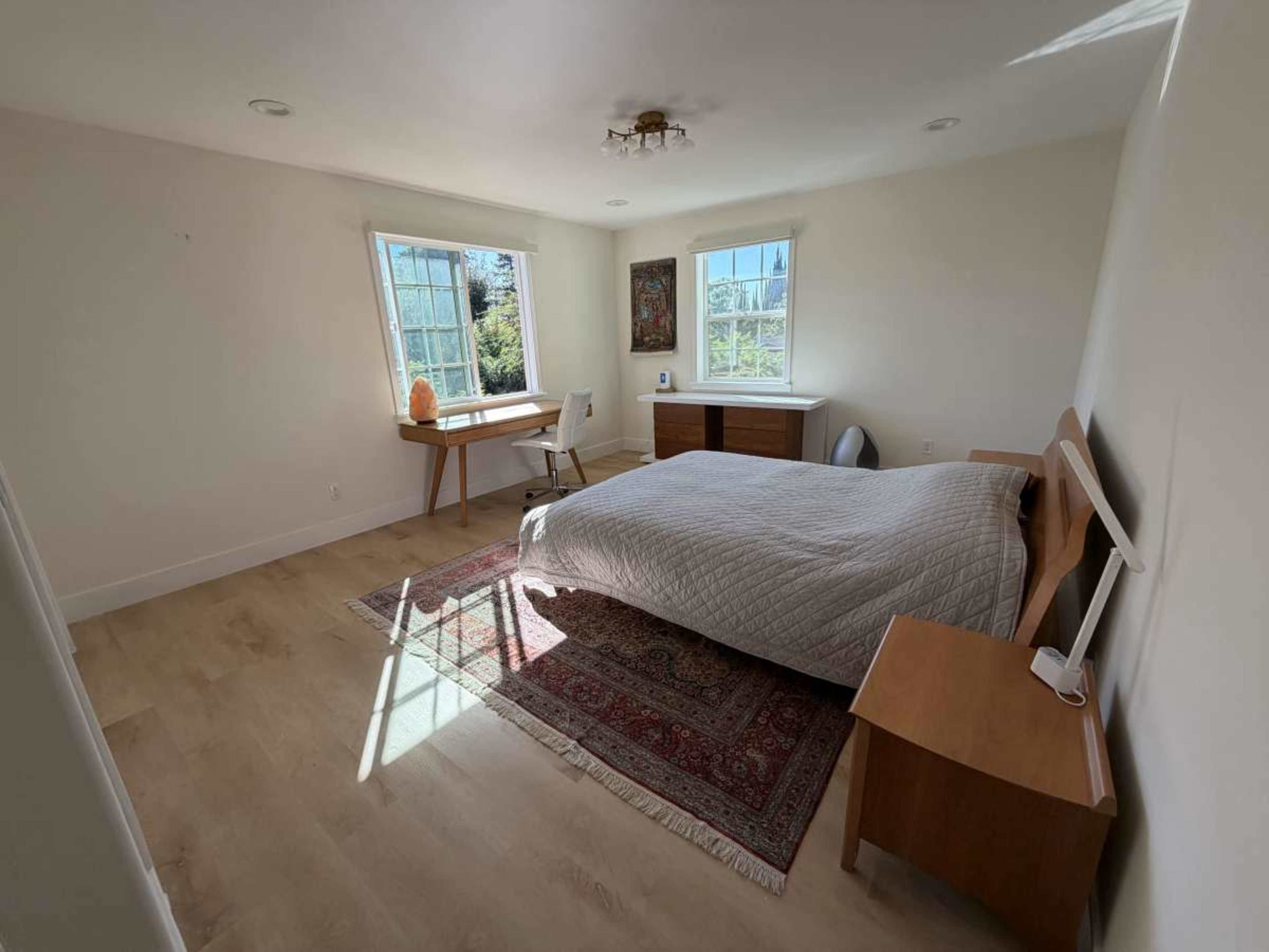 The image shows a well-lit bedroom featuring a bed with a quilt, a wooden desk and chair, and large windows allowing natural light to enter.