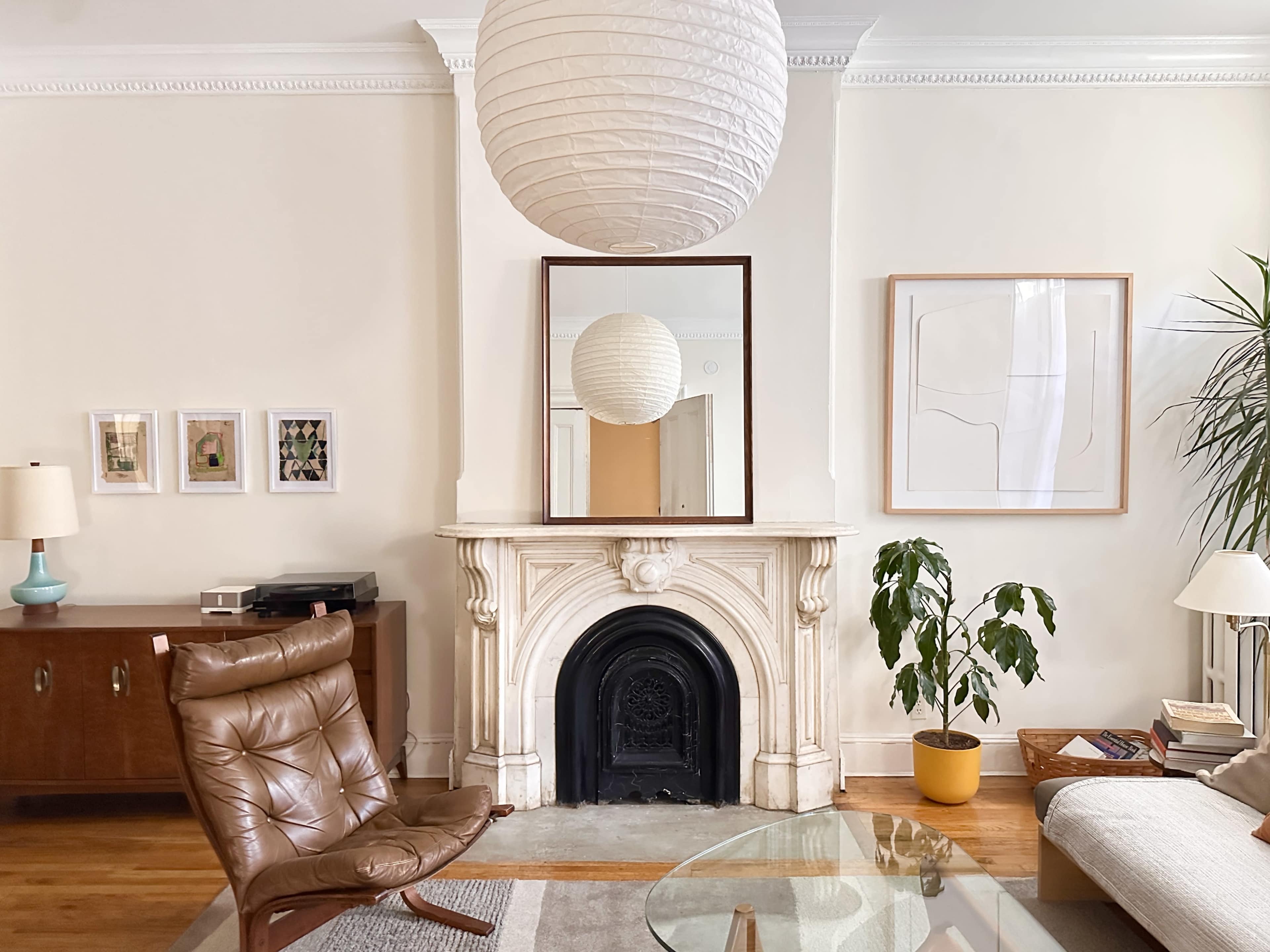 The image shows a living room featuring a brown leather chair, a modern lamp, a marble fireplace, a large round paper lantern, and decorative artwork on the walls.