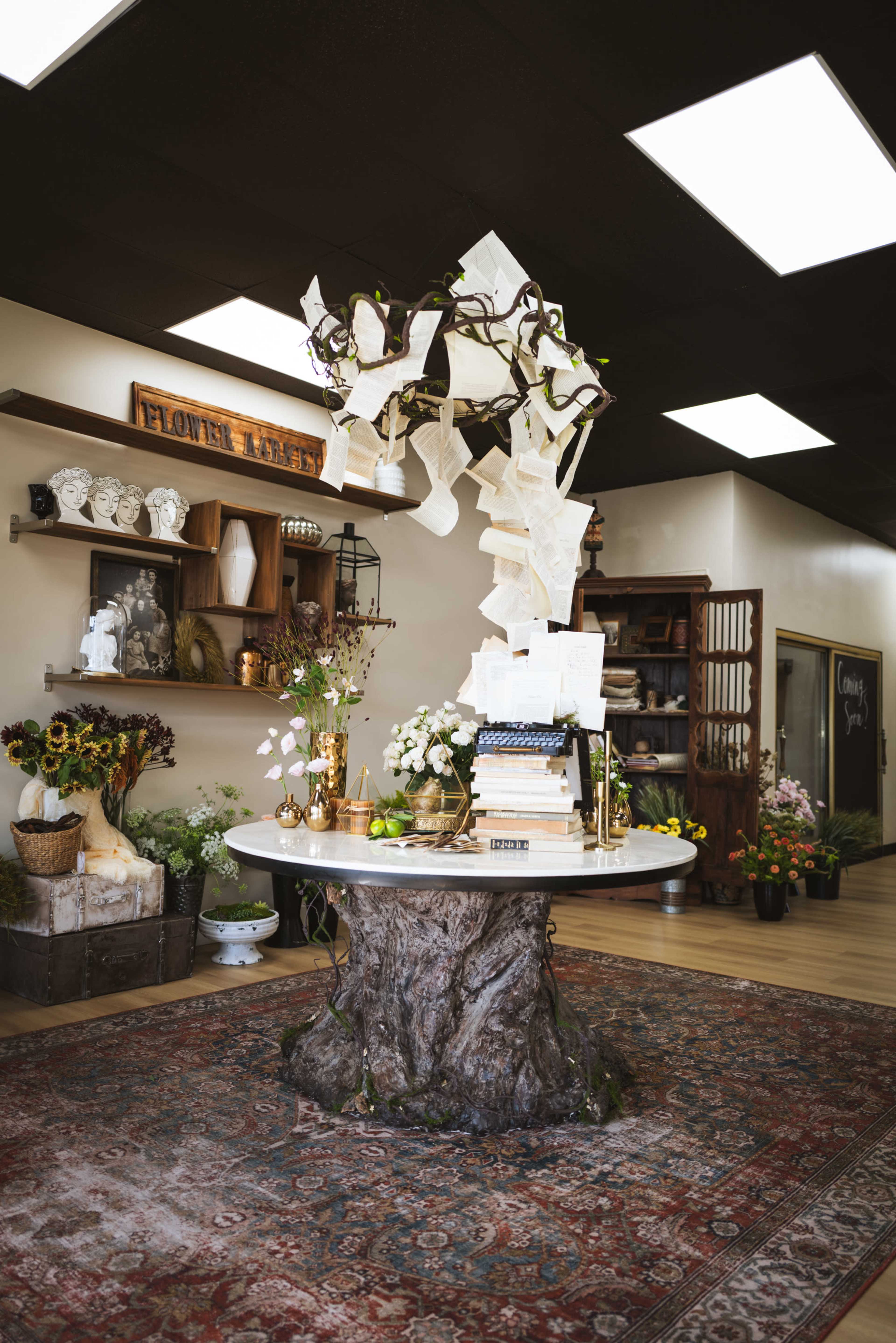 The image shows a flower shop interior with a decorative table made from a tree stump, surrounded by various plants and floral arrangements, under a ceiling with bright lights.