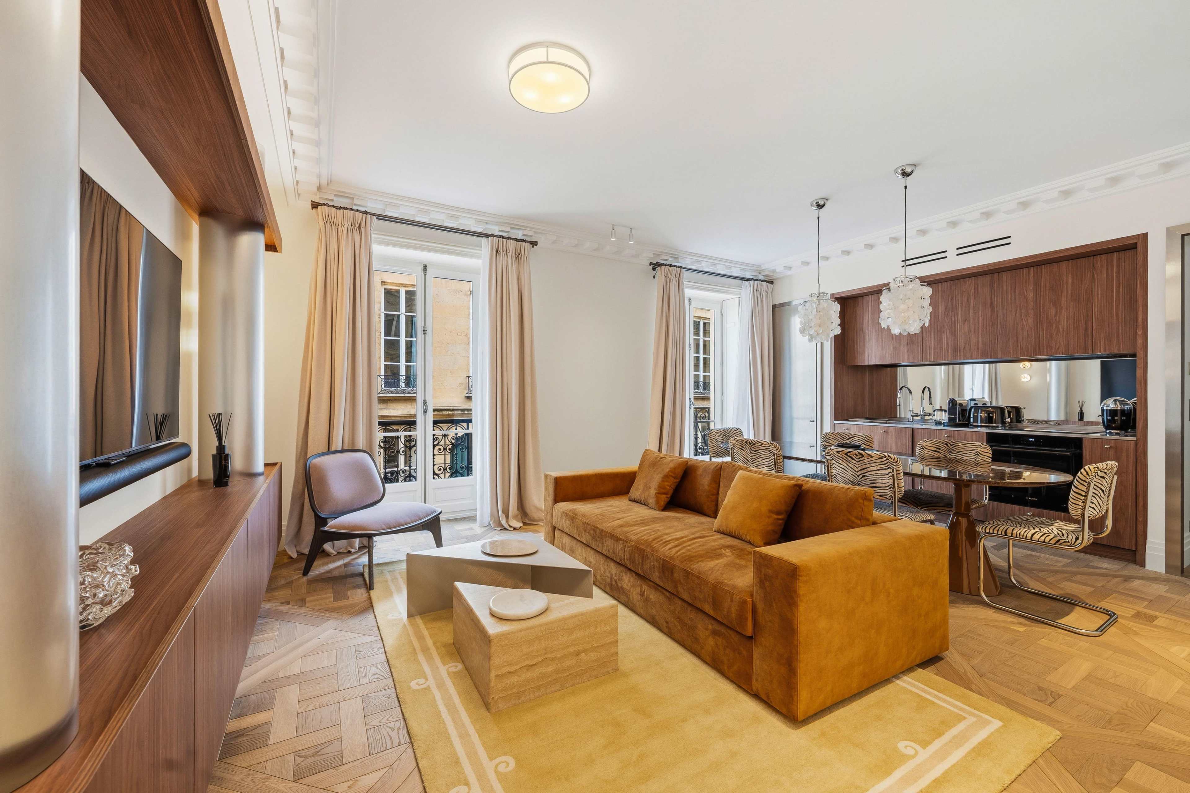 The image shows a modern living room featuring a brown sofa, a wooden entertainment unit, and a dining area with sleek chairs and a kitchen counter.