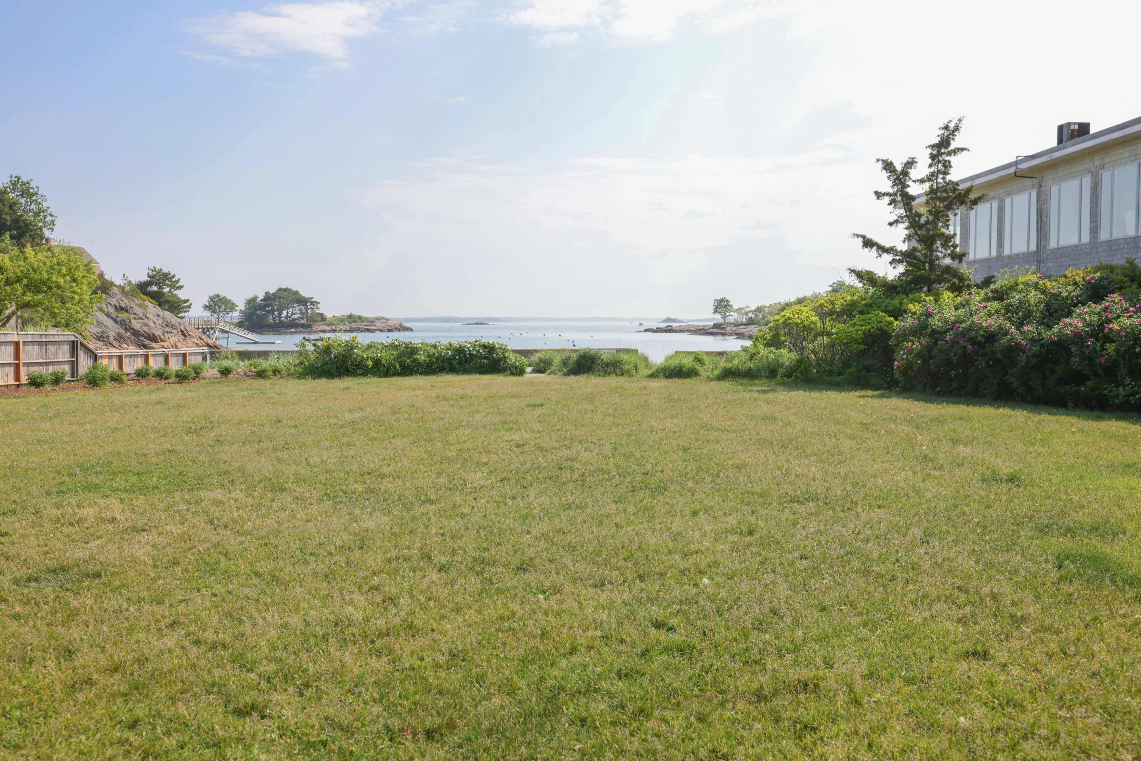 A wide, grassy field stretches towards a calm coastal bay with boats visible in the distance, framed by a house and trees on the right.