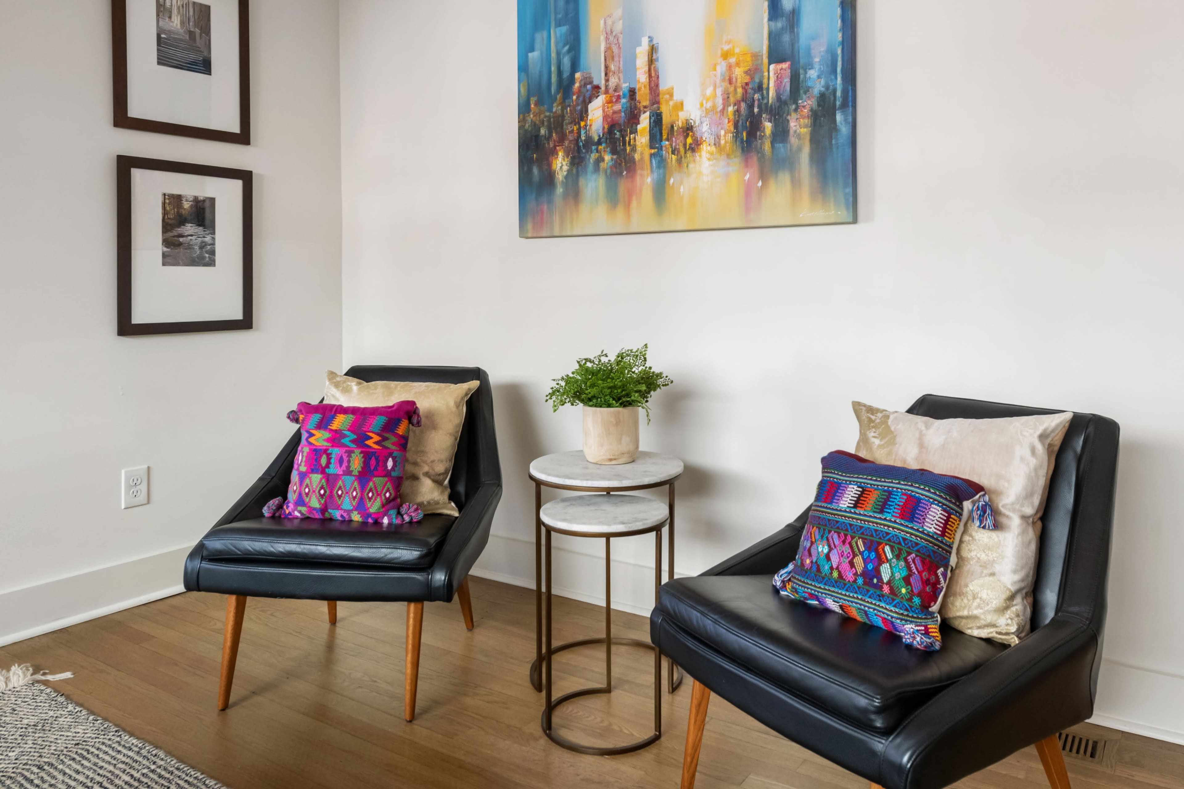 Two black lounge chairs with colorful pillows are positioned next to a small marble-top side table in a room featuring wall art and framed photographs.