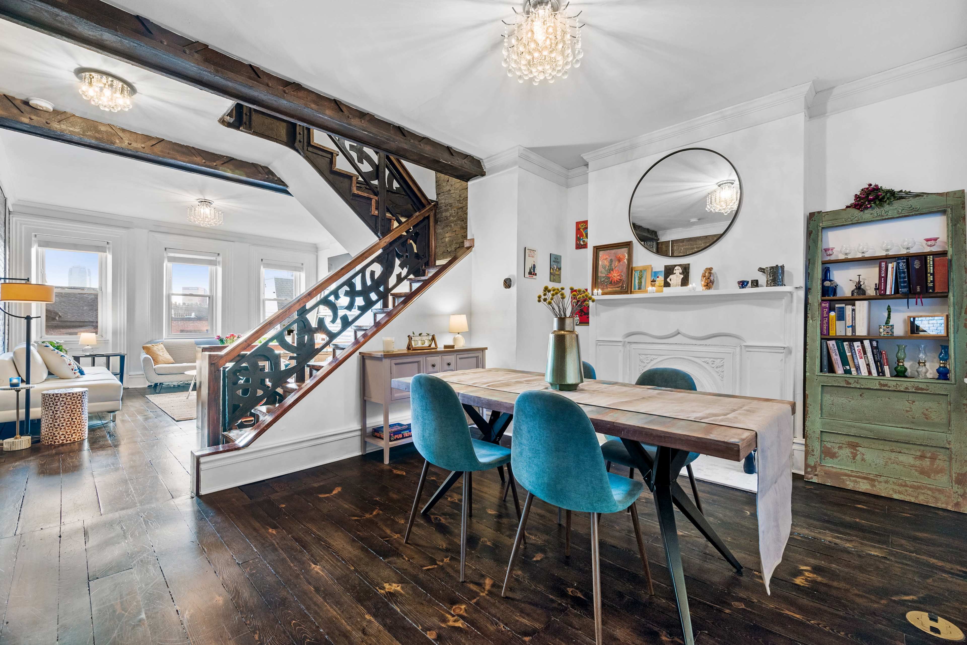 A well-decorated interior space featuring a wooden dining table, blue chairs, and a staircase with decorative railing, accompanied by various pieces of furniture and decor against a backdrop of large windows.