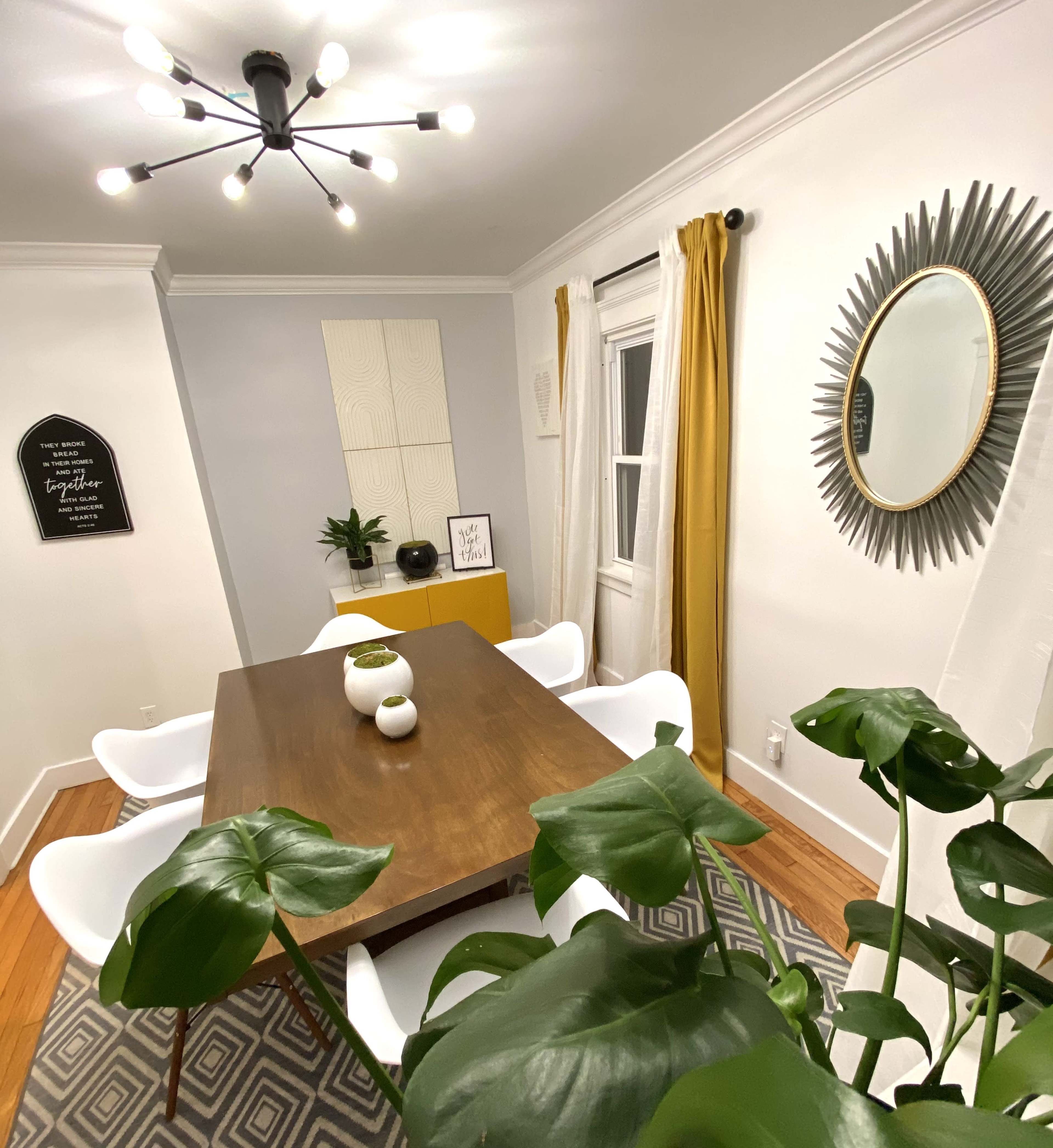 A dining room features a wooden table surrounded by white chairs, potted plants, and a decorative mirror on the wall.