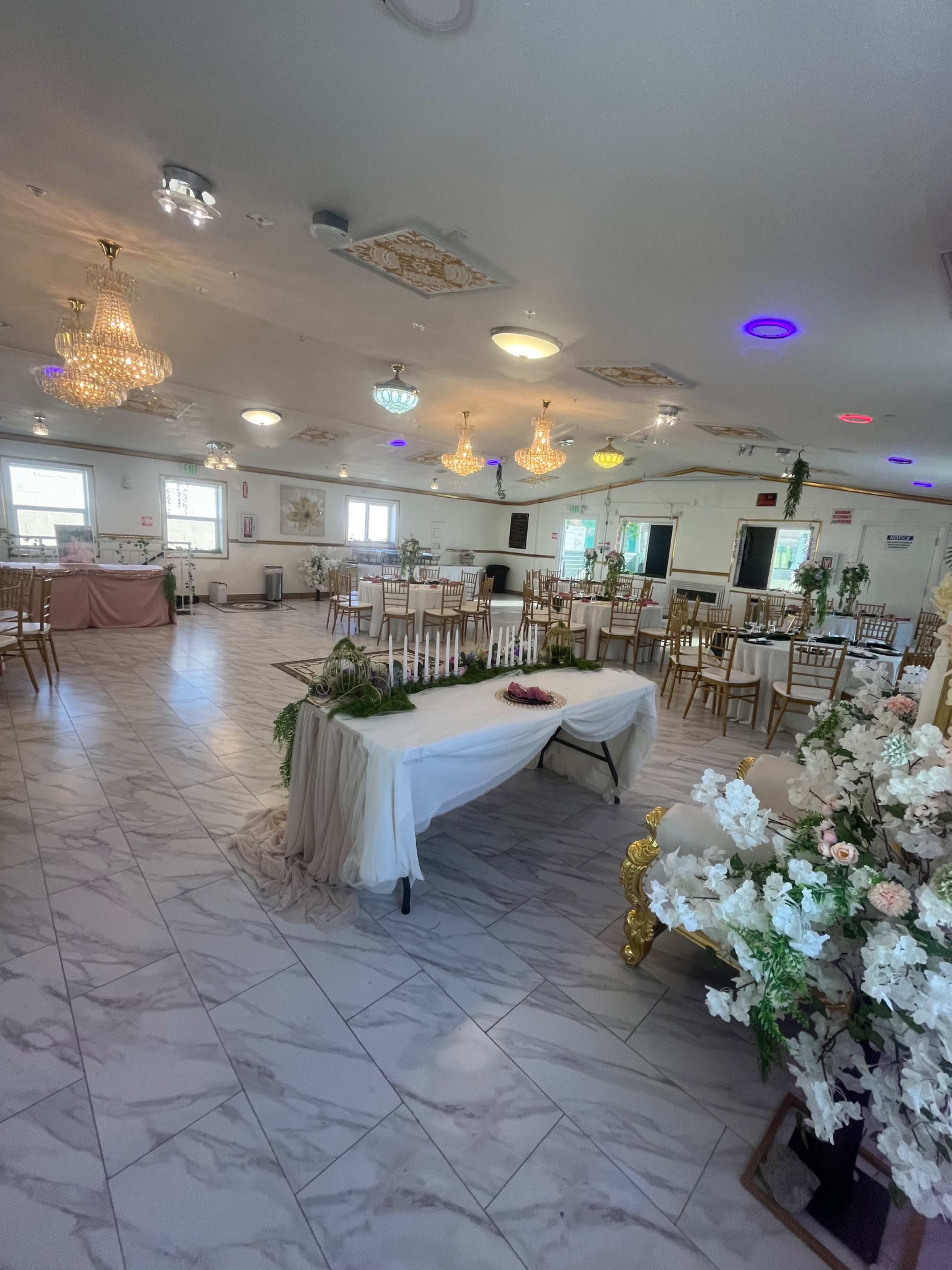 The image shows a decorated event hall with chandeliers, a long table at the front, and floral arrangements, set up for a gathering or celebration.