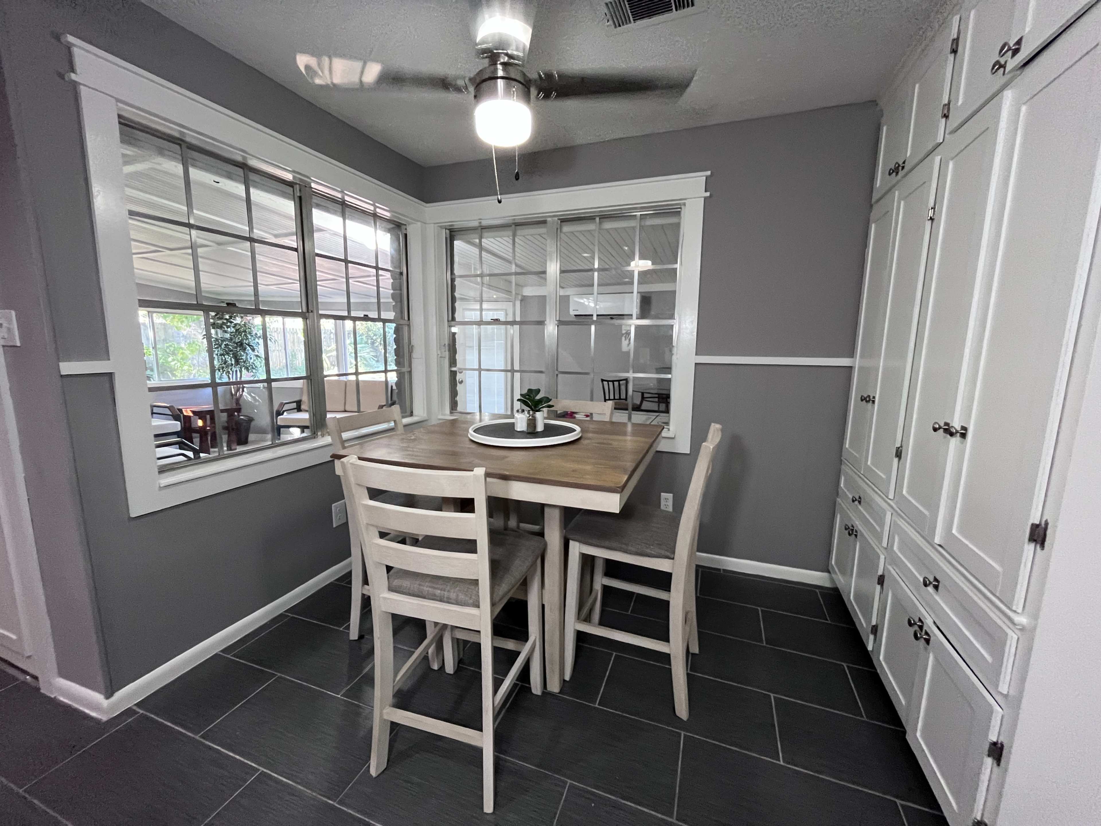 A dining area with a wooden table and four chairs is illuminated by a ceiling fan and features large windows that allow natural light to enter.