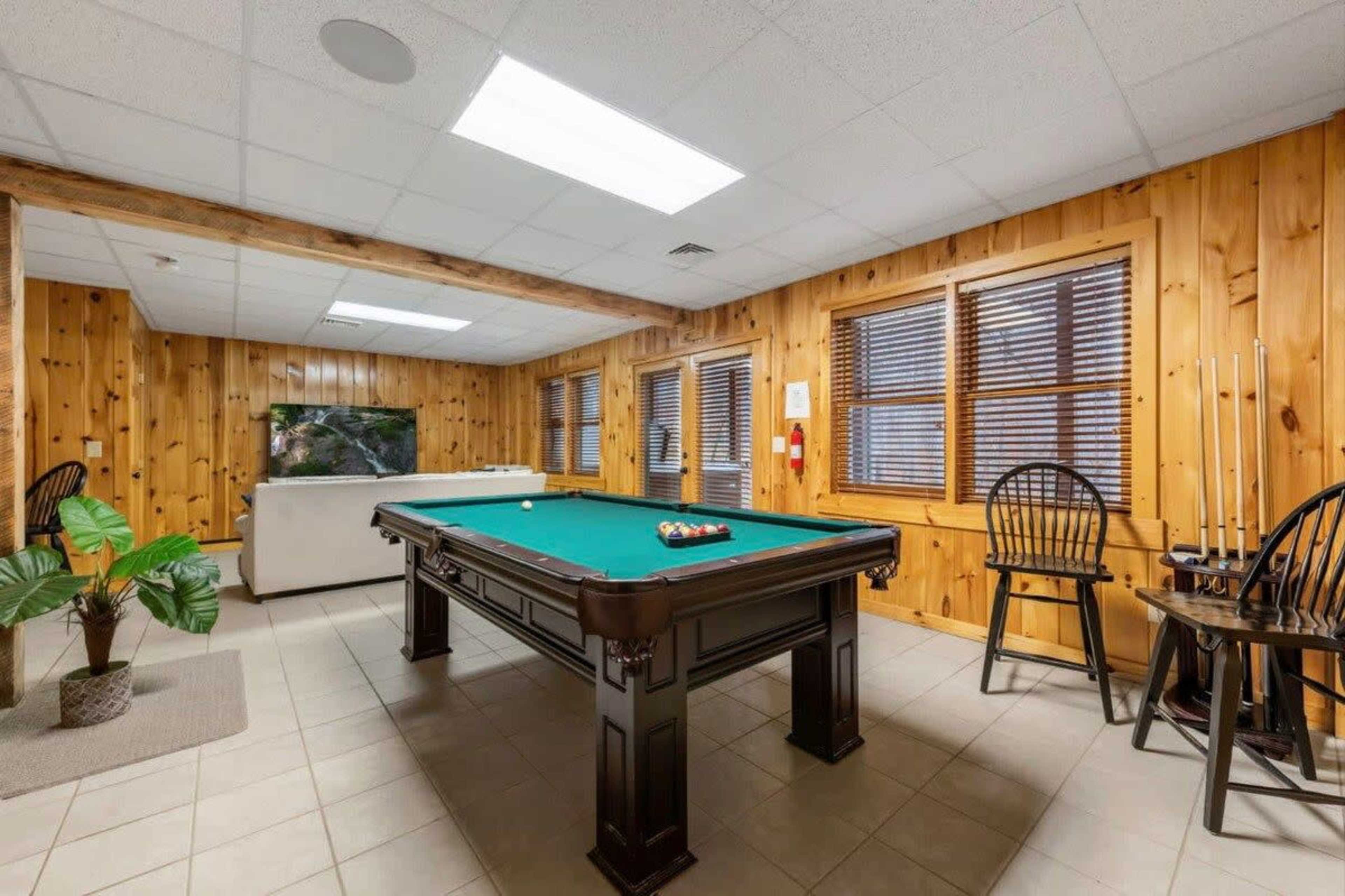 A game room featuring a billiard table in the center, surrounded by wooden-paneled walls and windows with blinds.