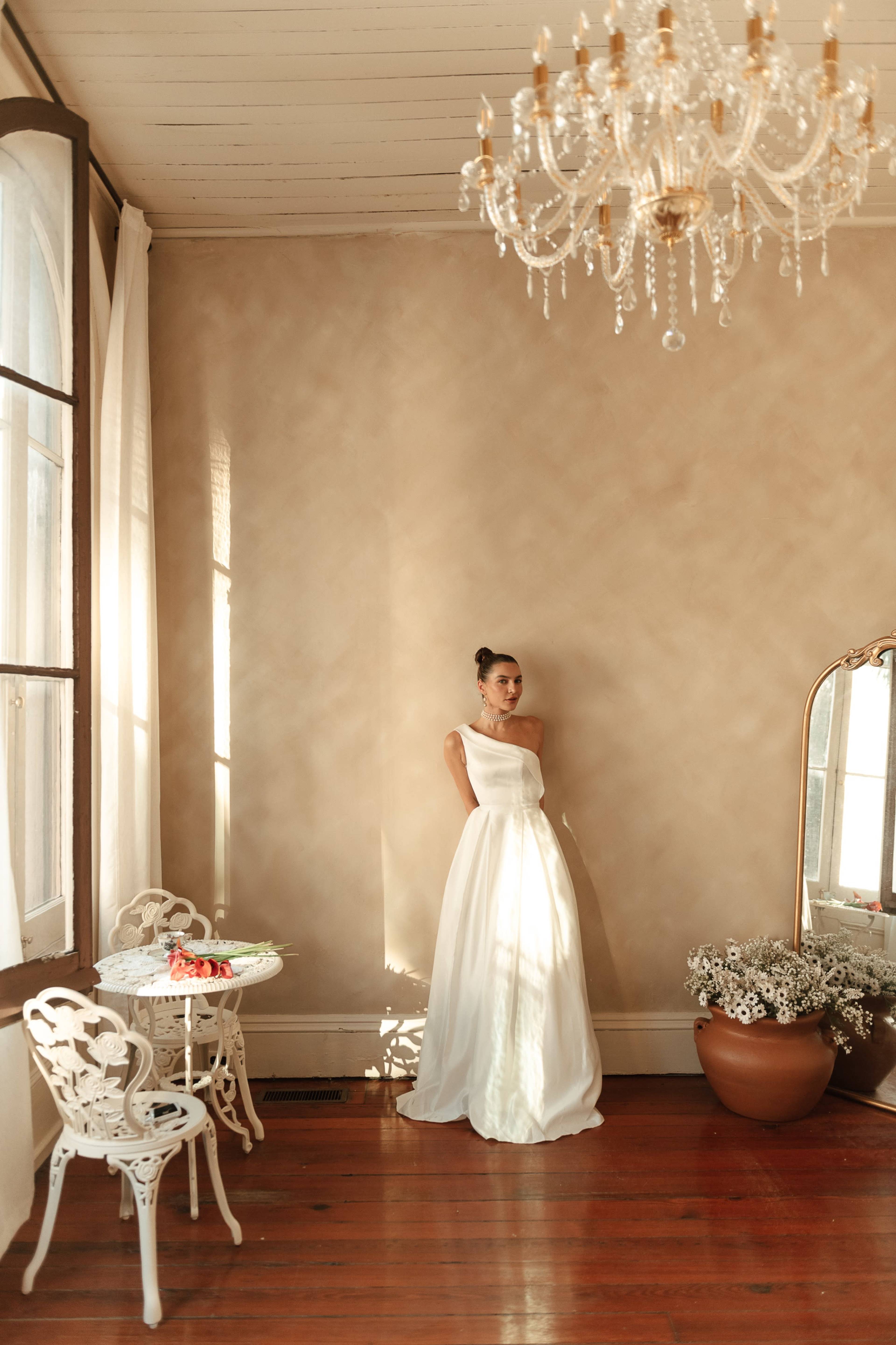A woman in a white dress stands beside a mirror in a sunlit room decorated with a chandelier and a small table.